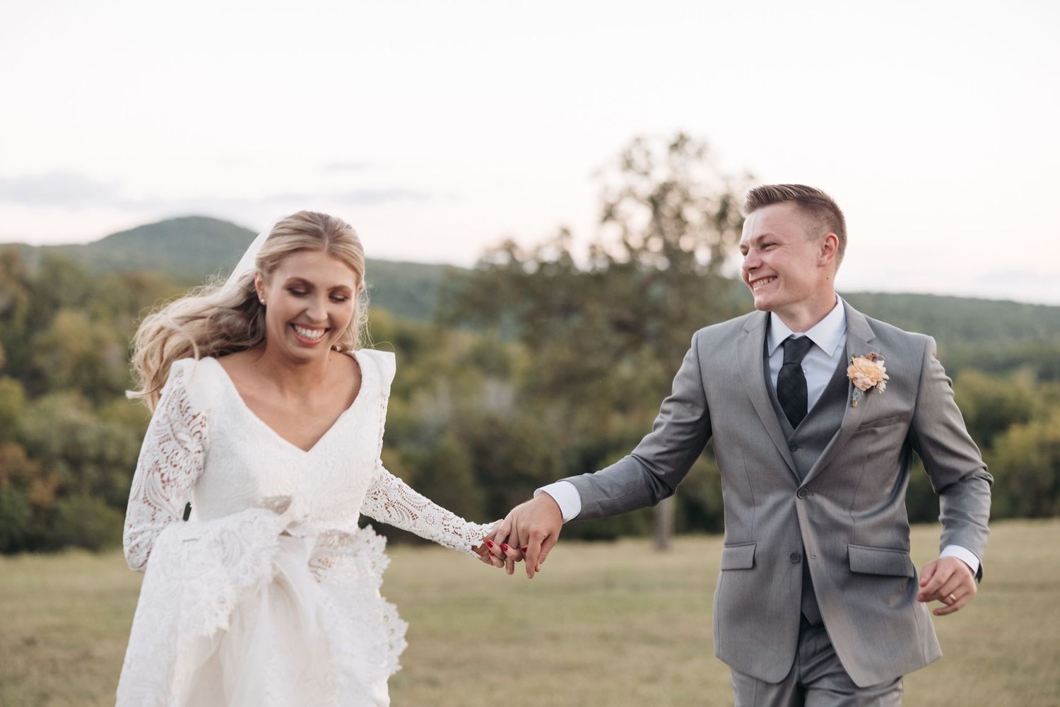 Happy newlyweds laughing while running through grassy field during sunset photo session.