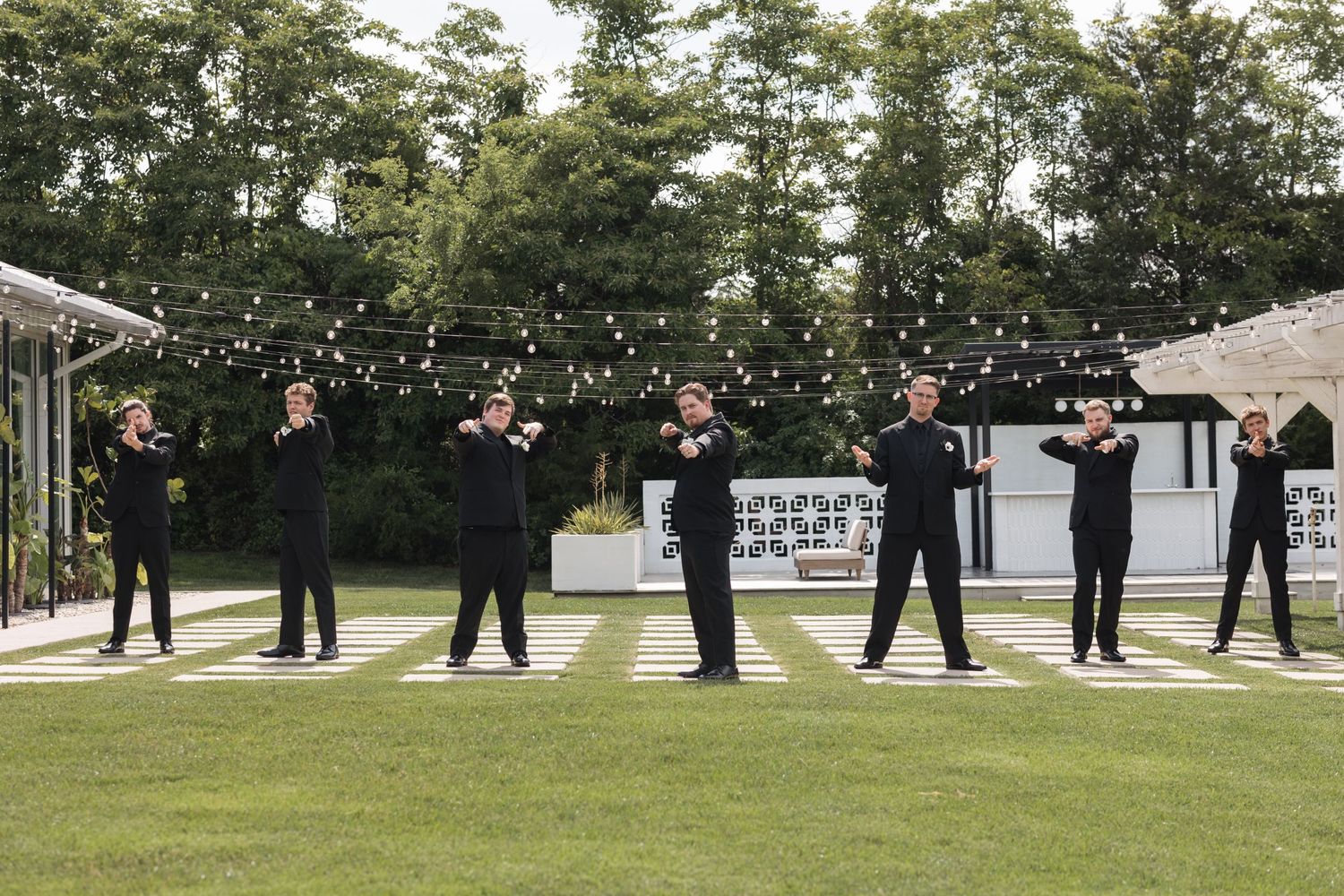 Wedding party members in black attire pose together on a lawn with string lights overhead.