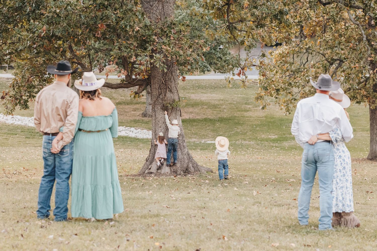 Family members watch young child playing in distance under autumn trees.