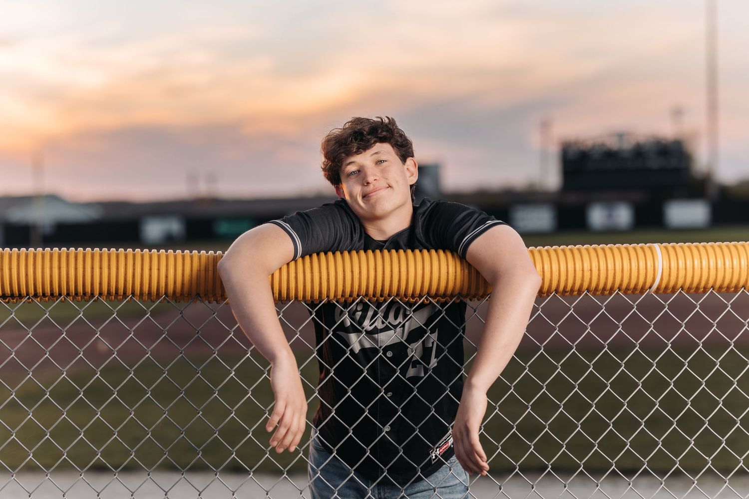 Athletic figure leaning on yellow fence during sunset at sports field.