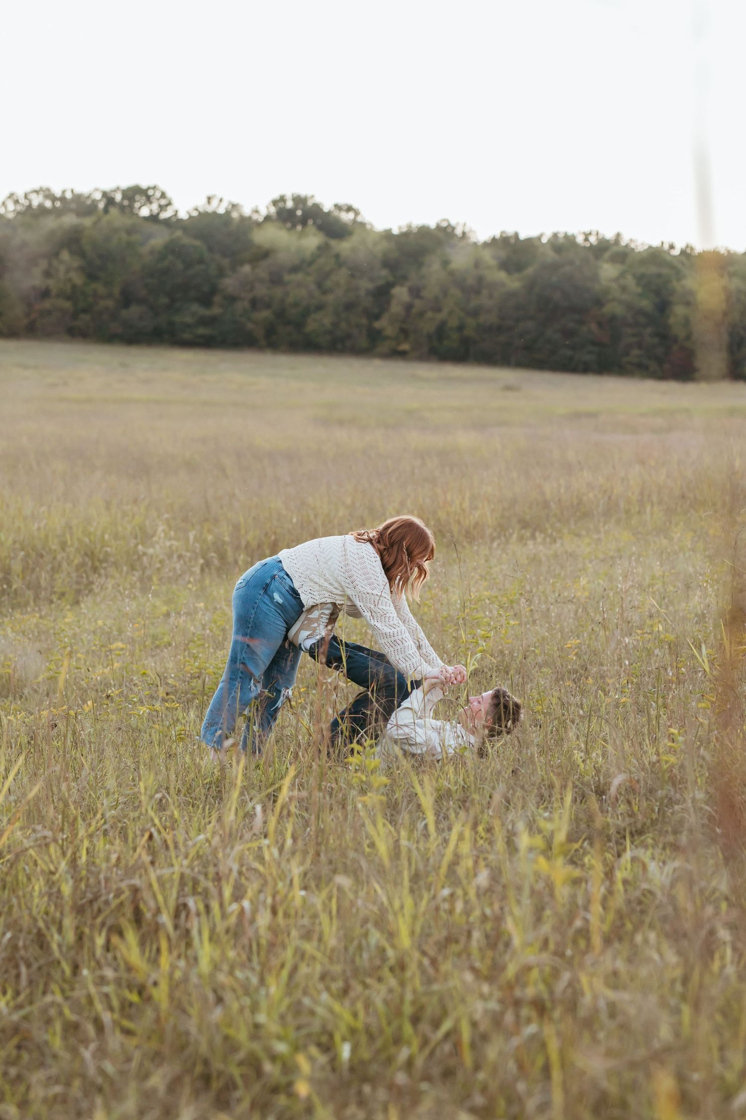 Person in white sweater and jeans bending down in tall grass field.