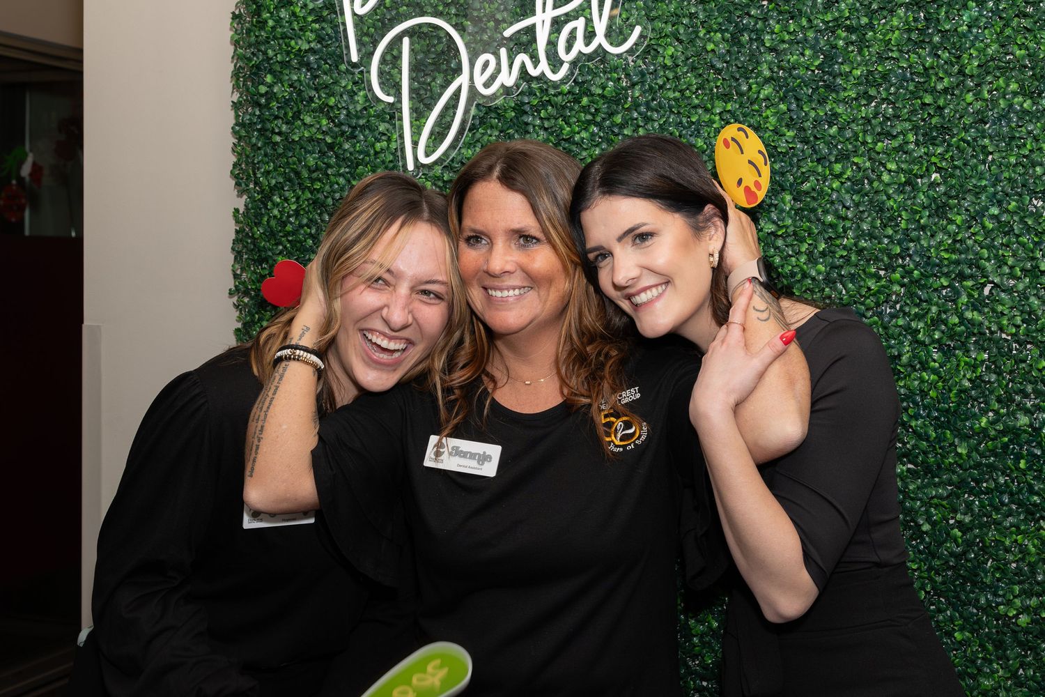 Three women pose together against a green hedge wall with a neon dental sign.