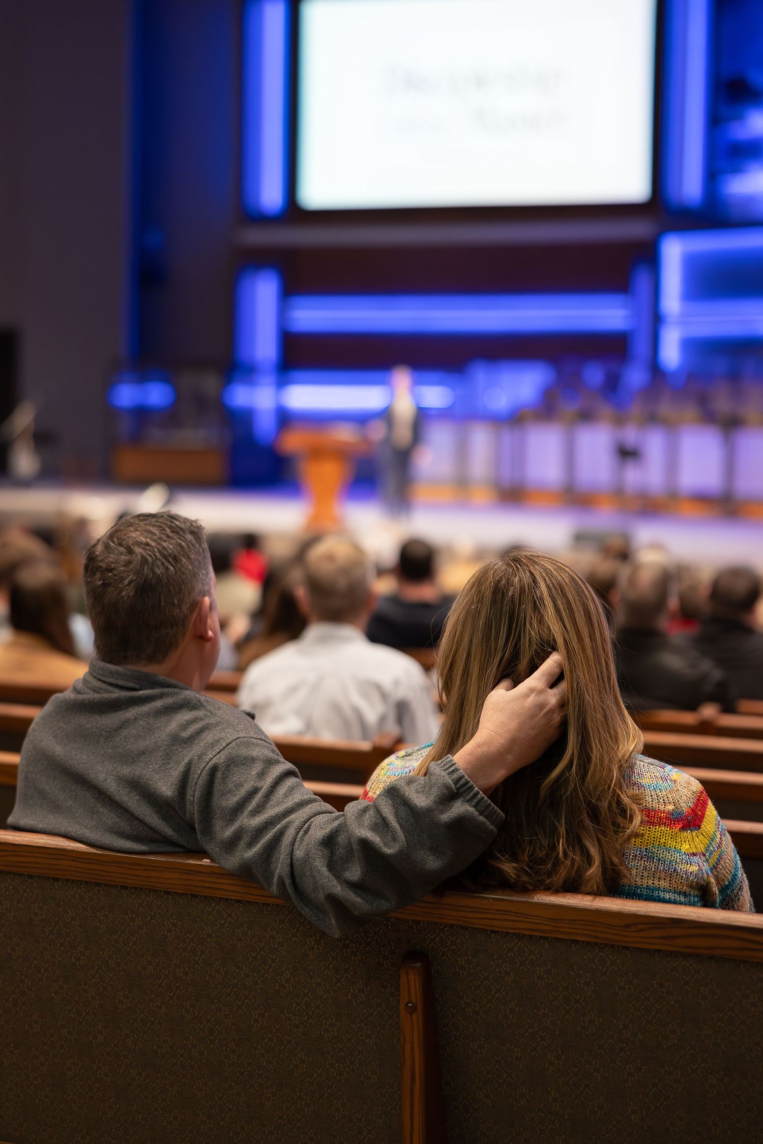 A couple sits together in church pews during a service with blue stage lighting visible.