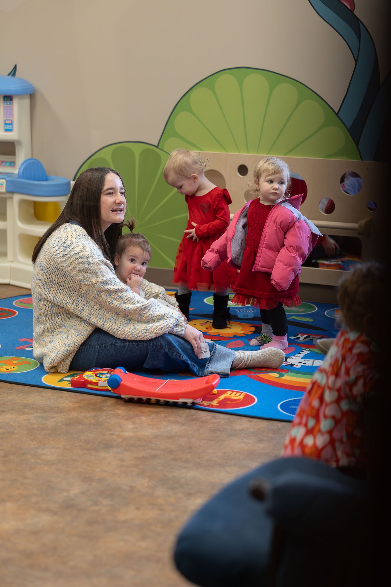 Playroom scene with colorful wall murals and children sitting on alphabet learning carpet.
