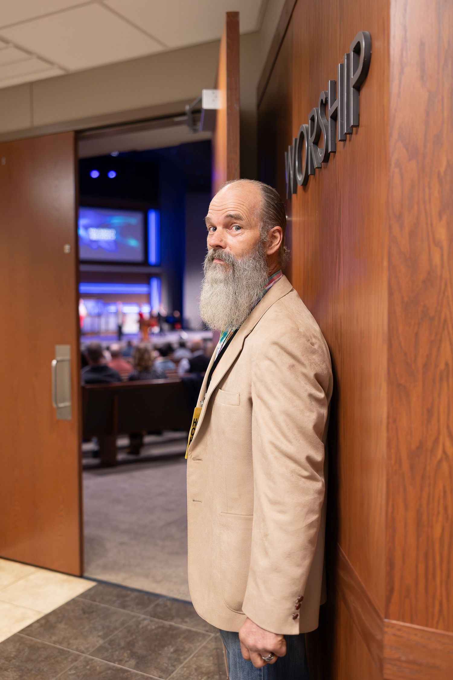 A man with a long beard wearing a tan blazer stands in a hallway.