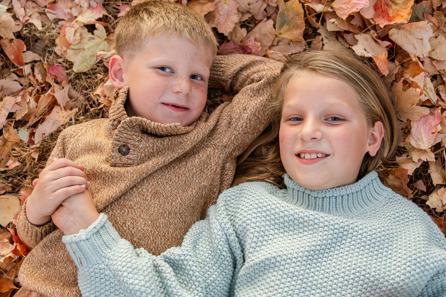 Two children lying in autumn leaves wearing cozy sweaters.