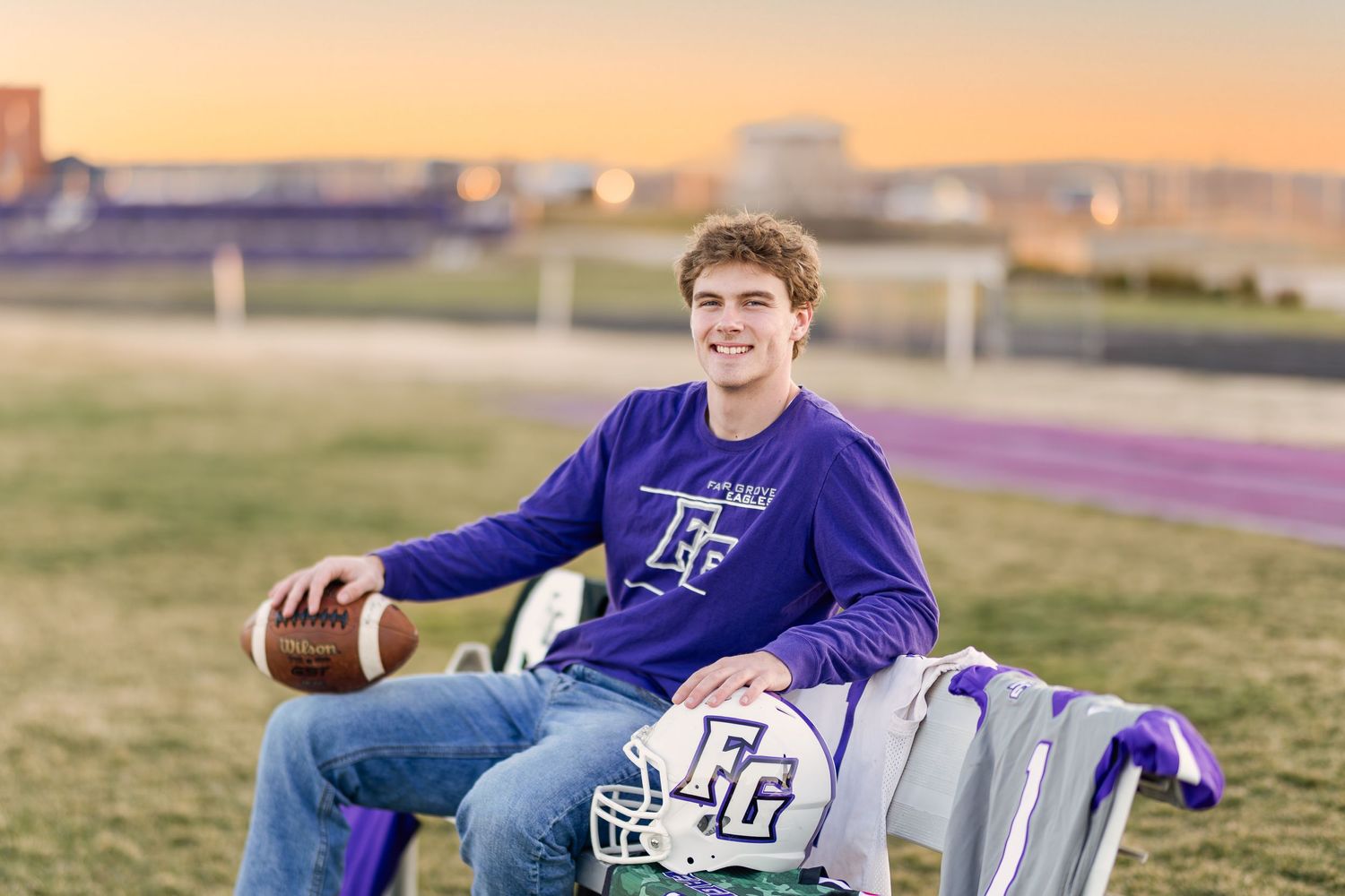 High school football player in purple team jersey sits on sideline with helmet and football at sunset.