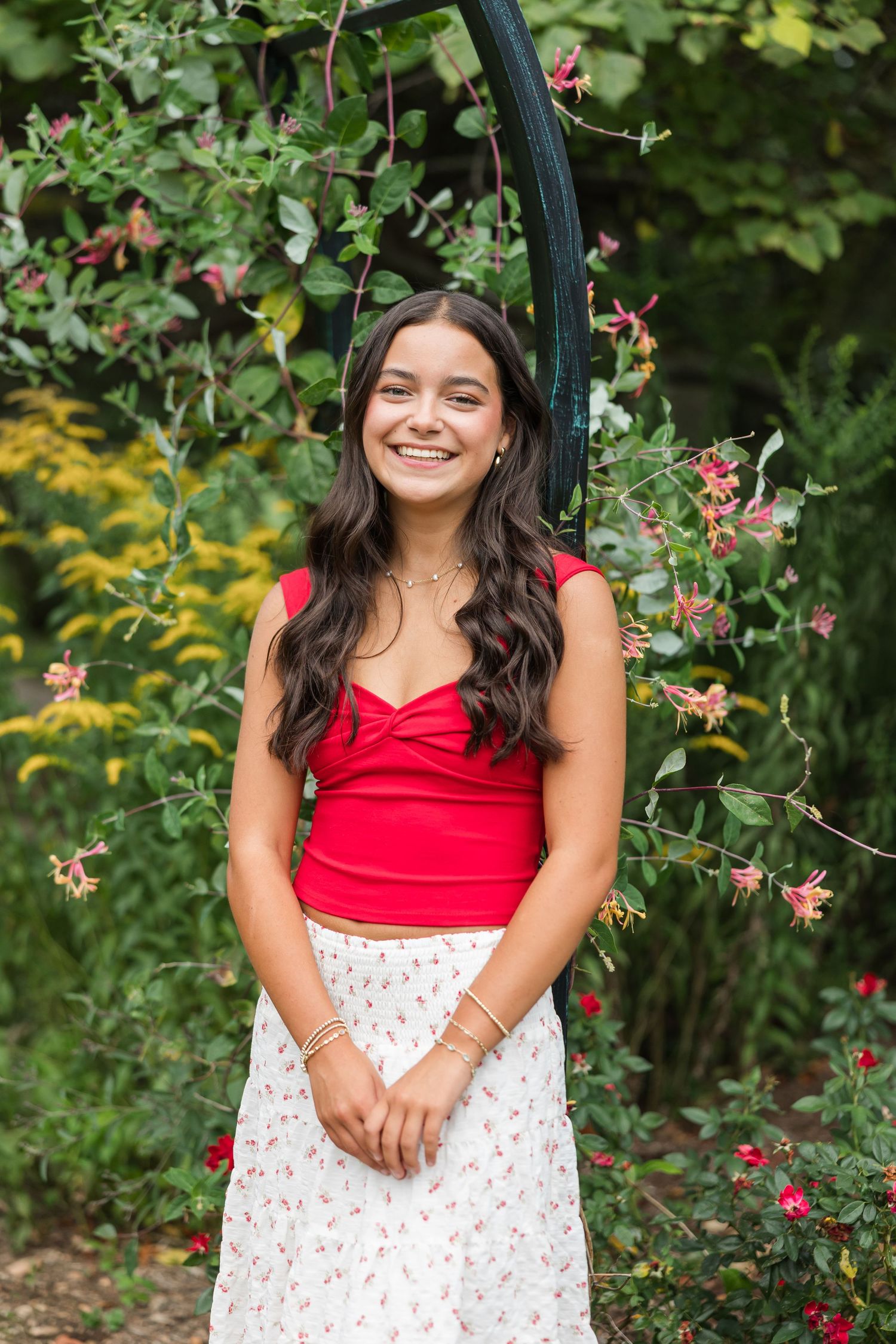 A person wearing a red top and floral skirt poses outdoors in a garden with blooming flowers and greenery.