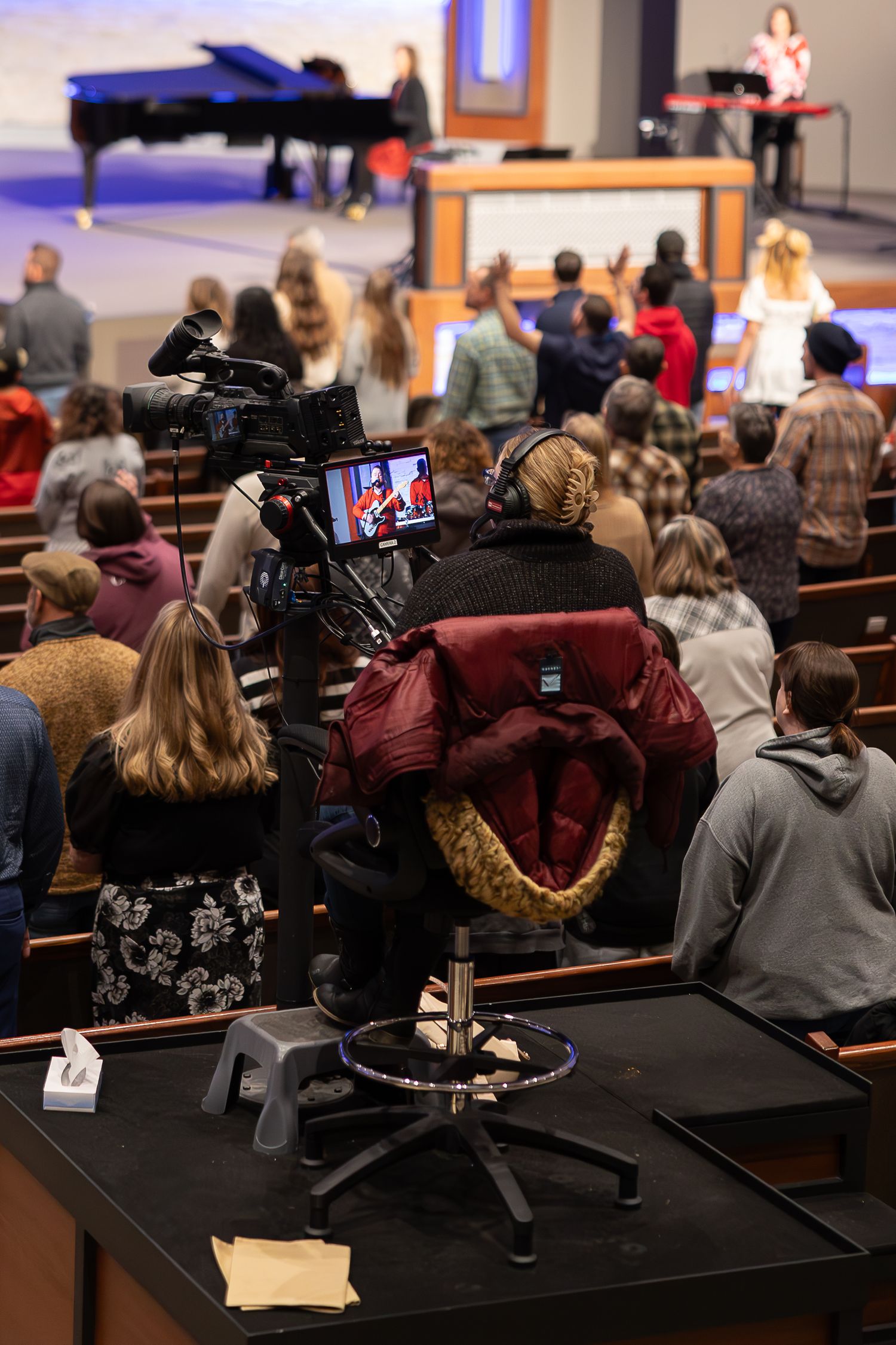 Professional video camera setup recording an event in an auditorium with piano and audience.