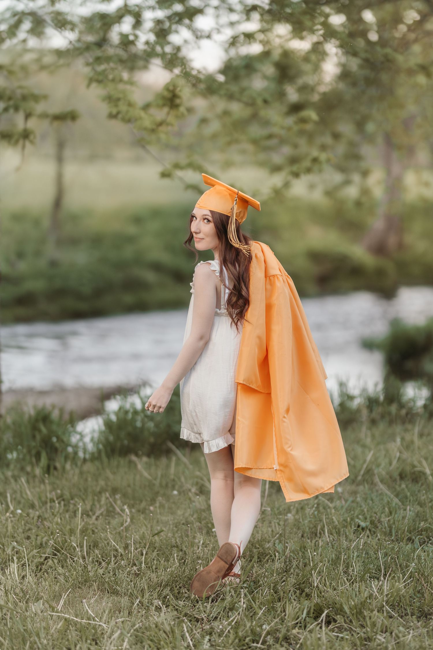 A graduate in orange cap and gown poses playfully in a grassy area near a stream.