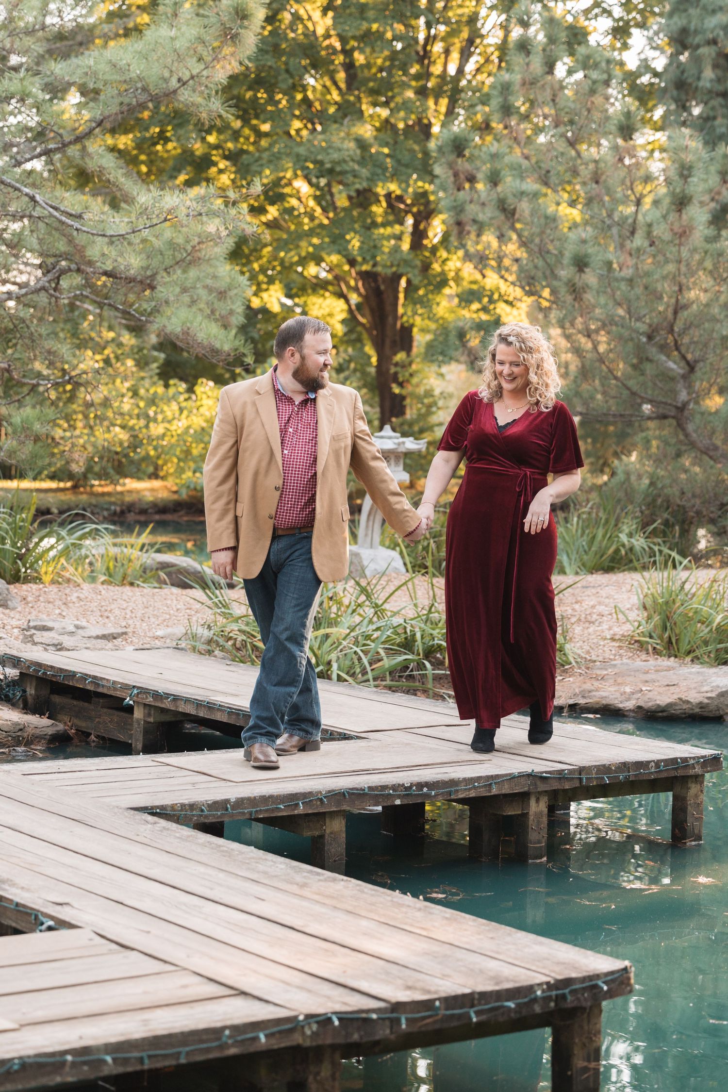 A couple walks hand in hand on a wooden dock over turquoise water surrounded by autumn foliage.