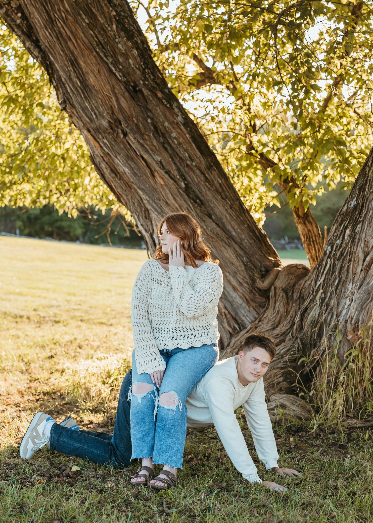 Playful couple creates fun poses around a large tree trunk in a sunny field.