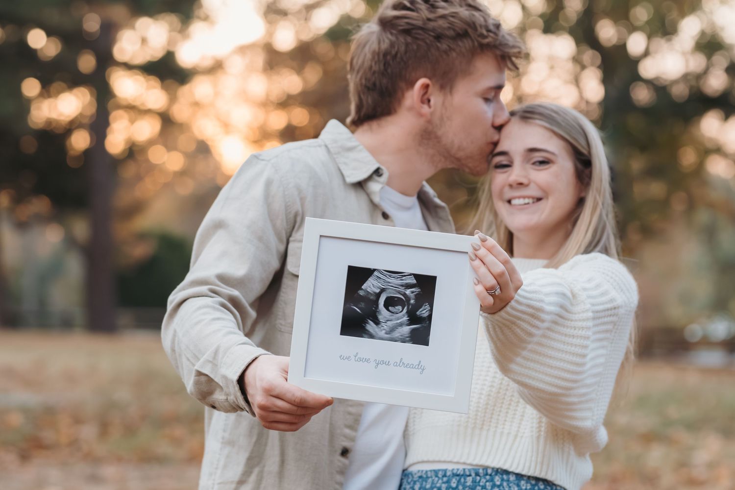 Happy couple displaying framed ultrasound photo during autumn photoshoot.