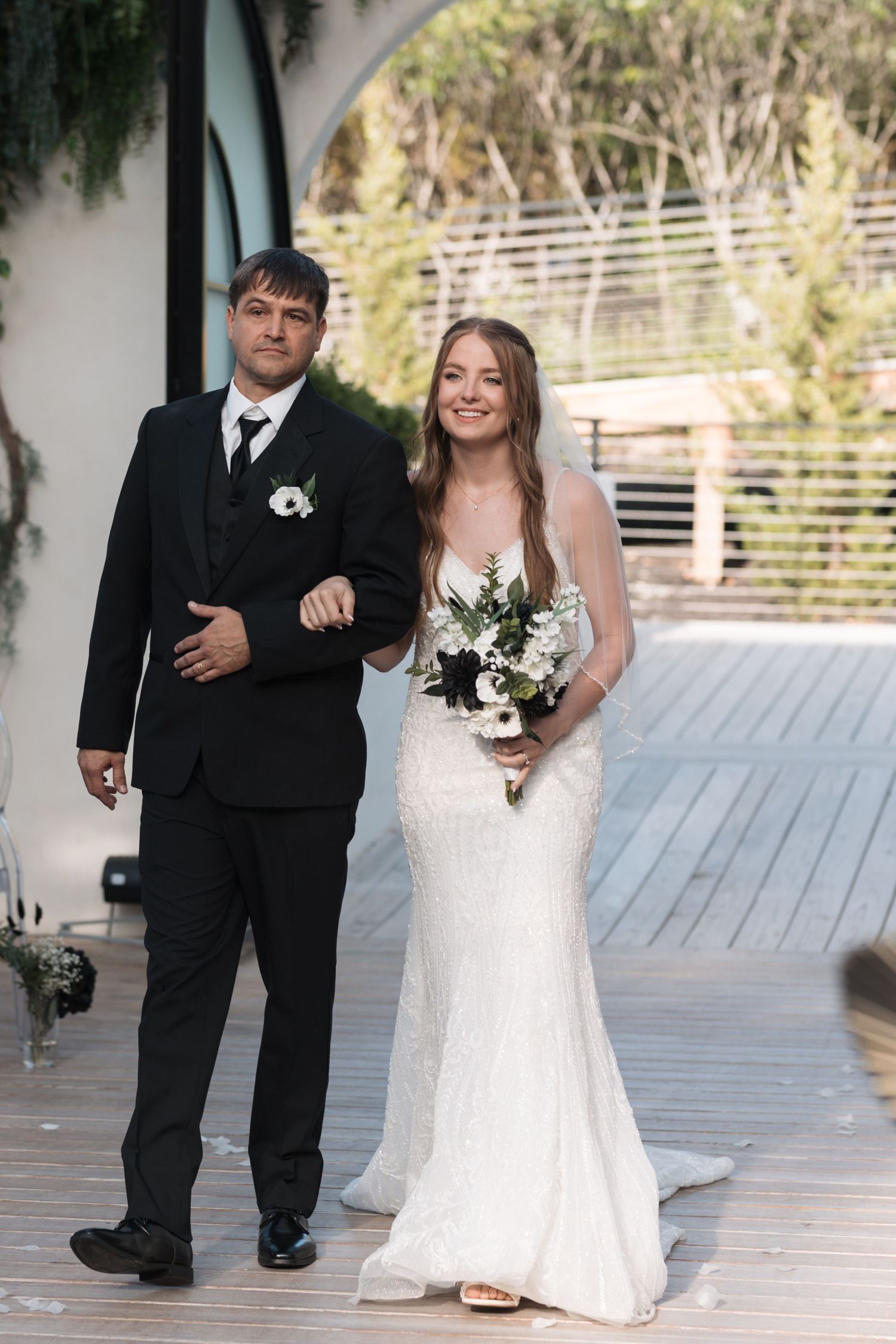 A wedding couple walks together on a covered porch decorated with greenery.