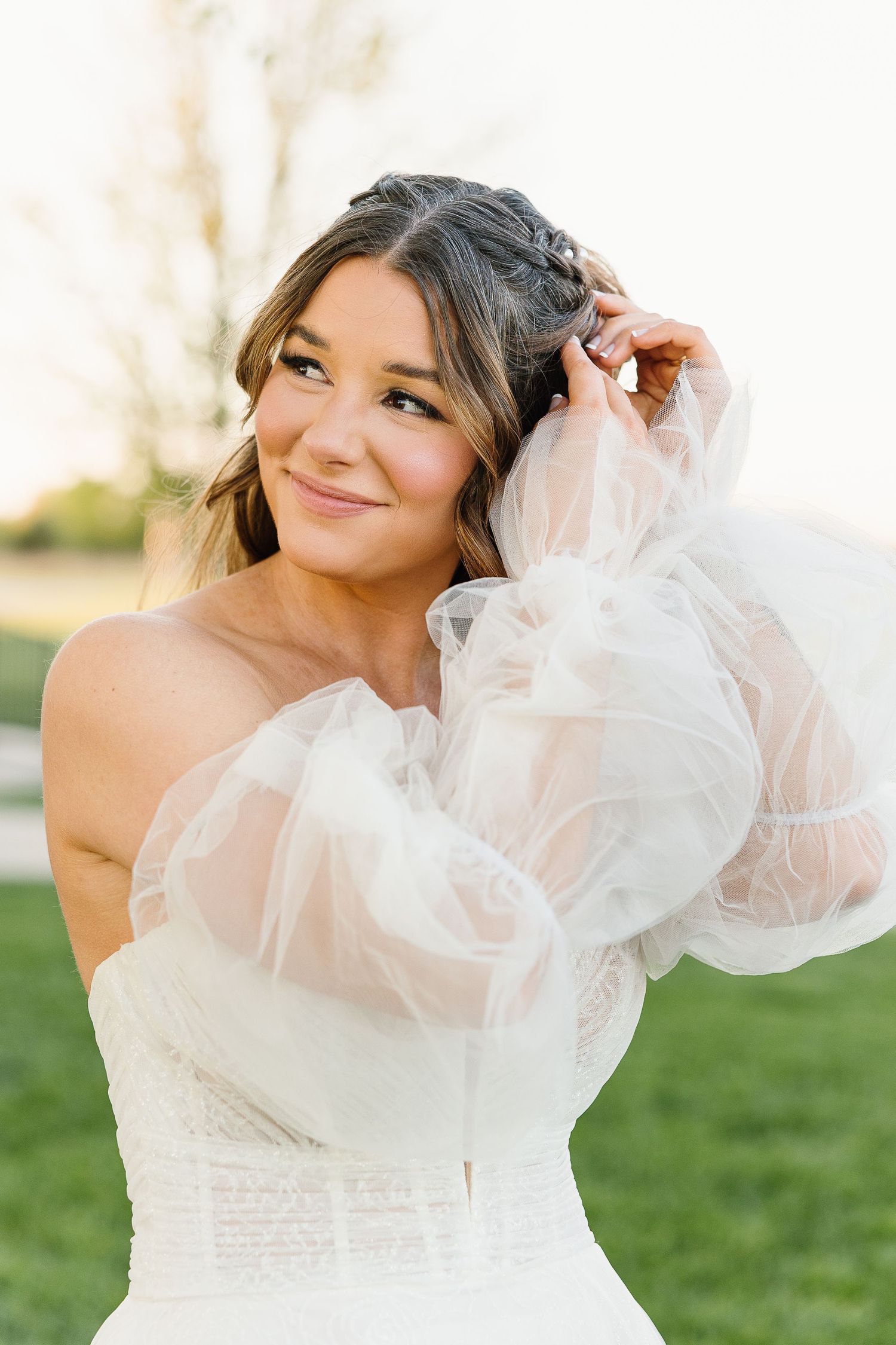 Bride in strapless wedding gown with sheer puff sleeves poses outdoors on sunny day.