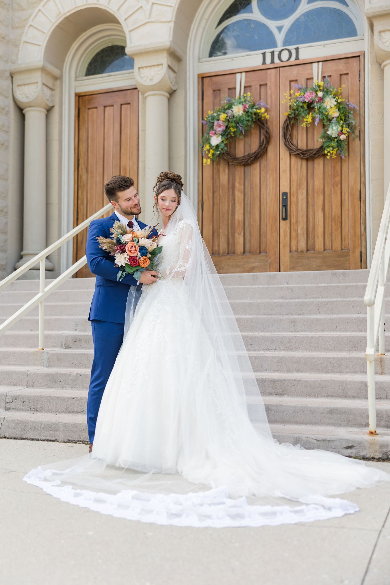 Bride and groom pose on church steps, she holds colorful bouquet, wearing white gown with long veil.