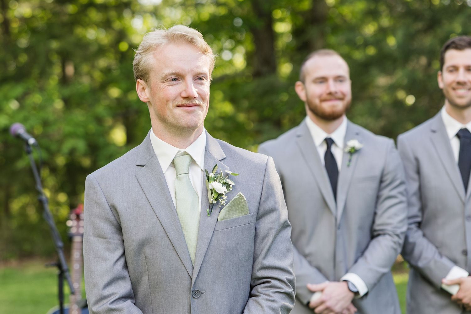 Groom in gray suit with boutonniere smiling at altar during outdoor wedding ceremony with groomsmen behind him.