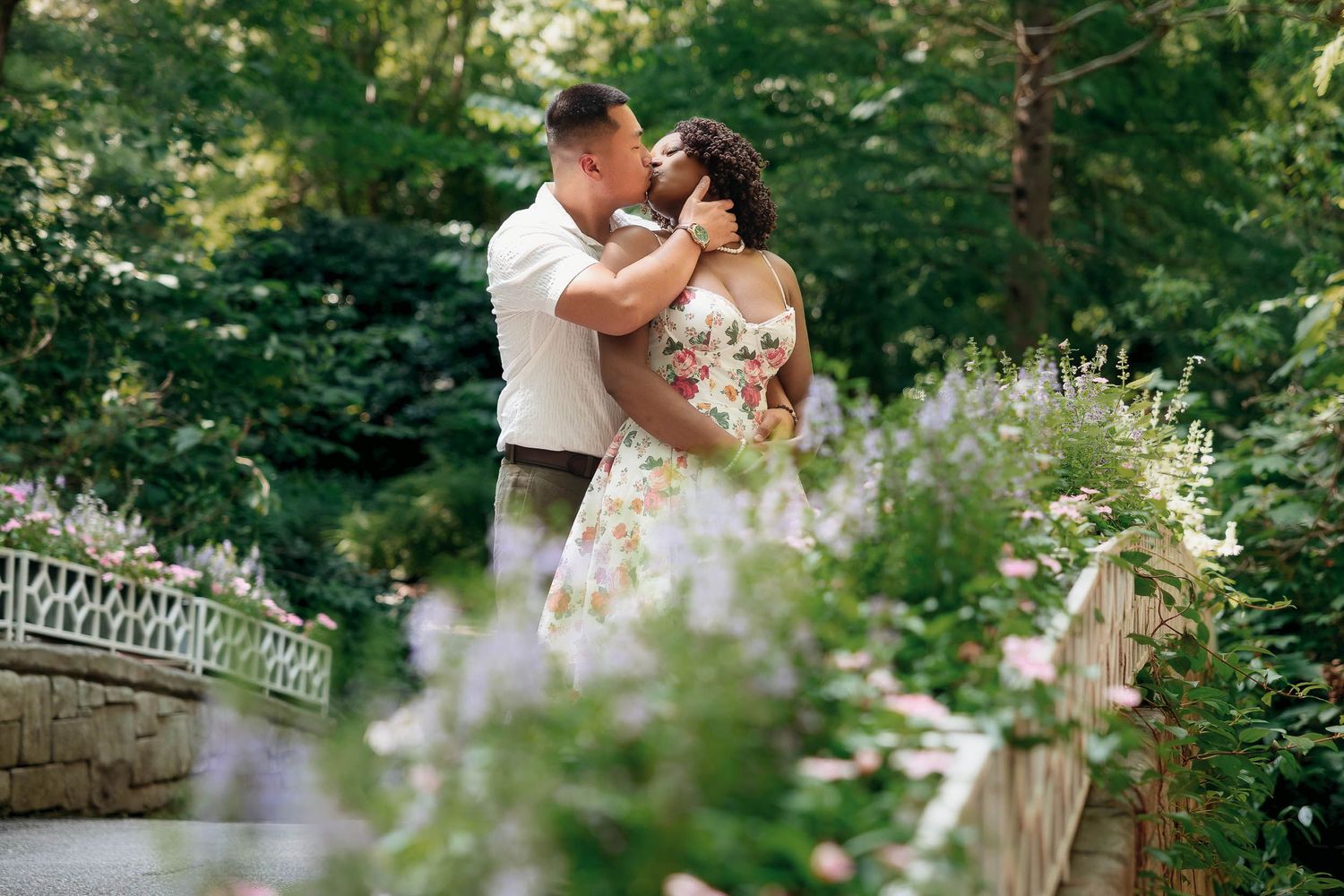 A romantic couple embraces surrounded by purple wildflowers in a garden setting with a decorative stone railing.