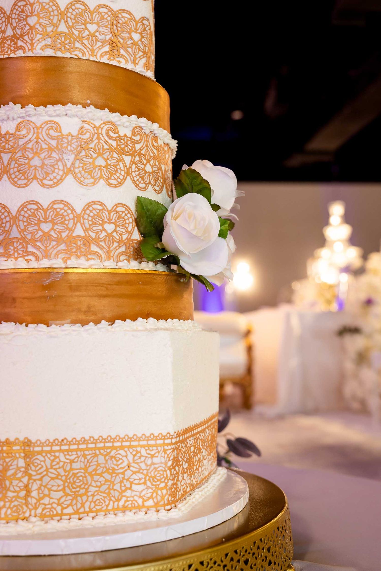Elegant four-tier white wedding cake with gold lace details and white roses on a gold stand at a reception.