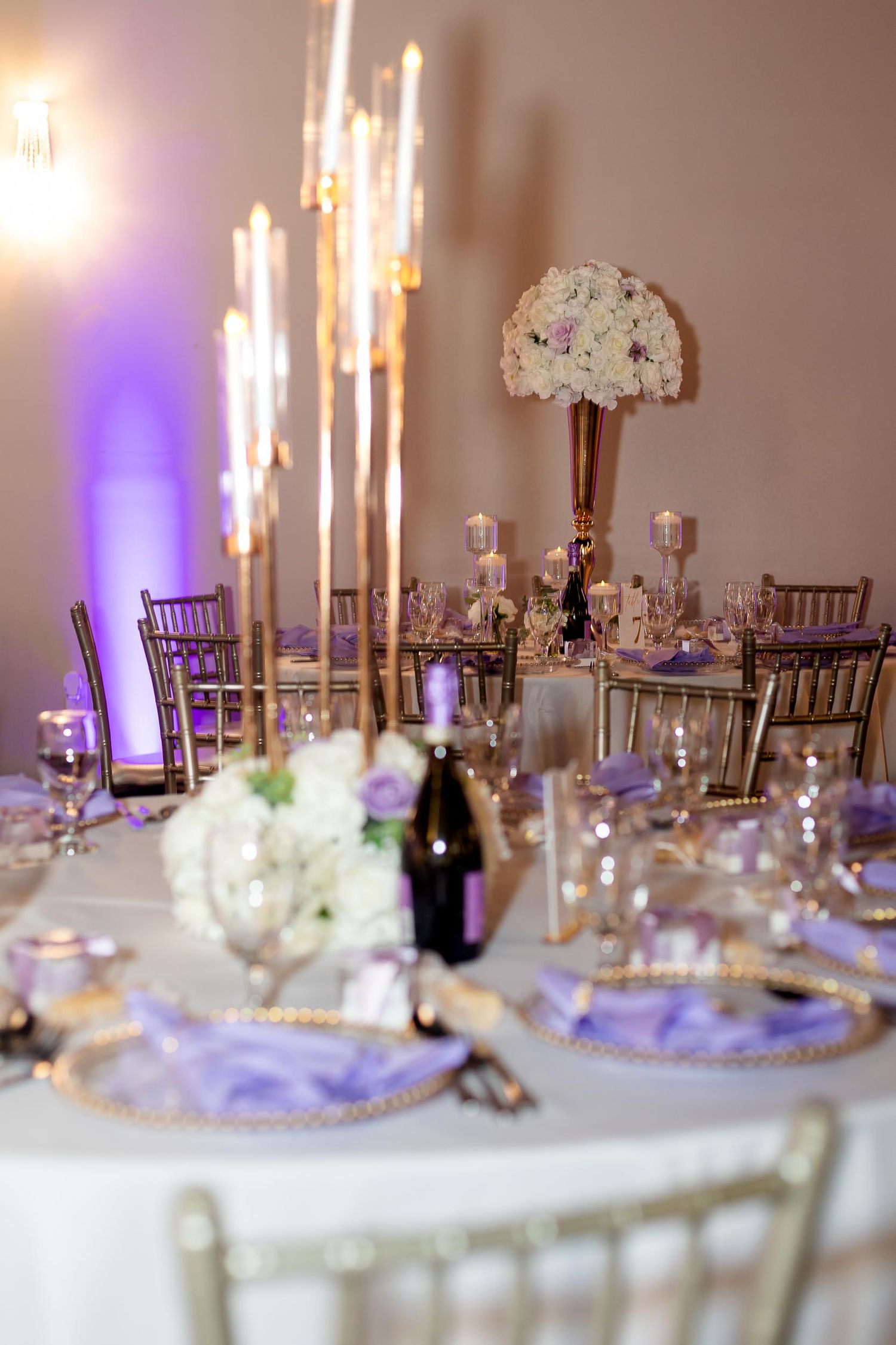Elegant wedding reception table with gold candelabras, purple charger plates, white floral centerpieces, and lavender uplighting.