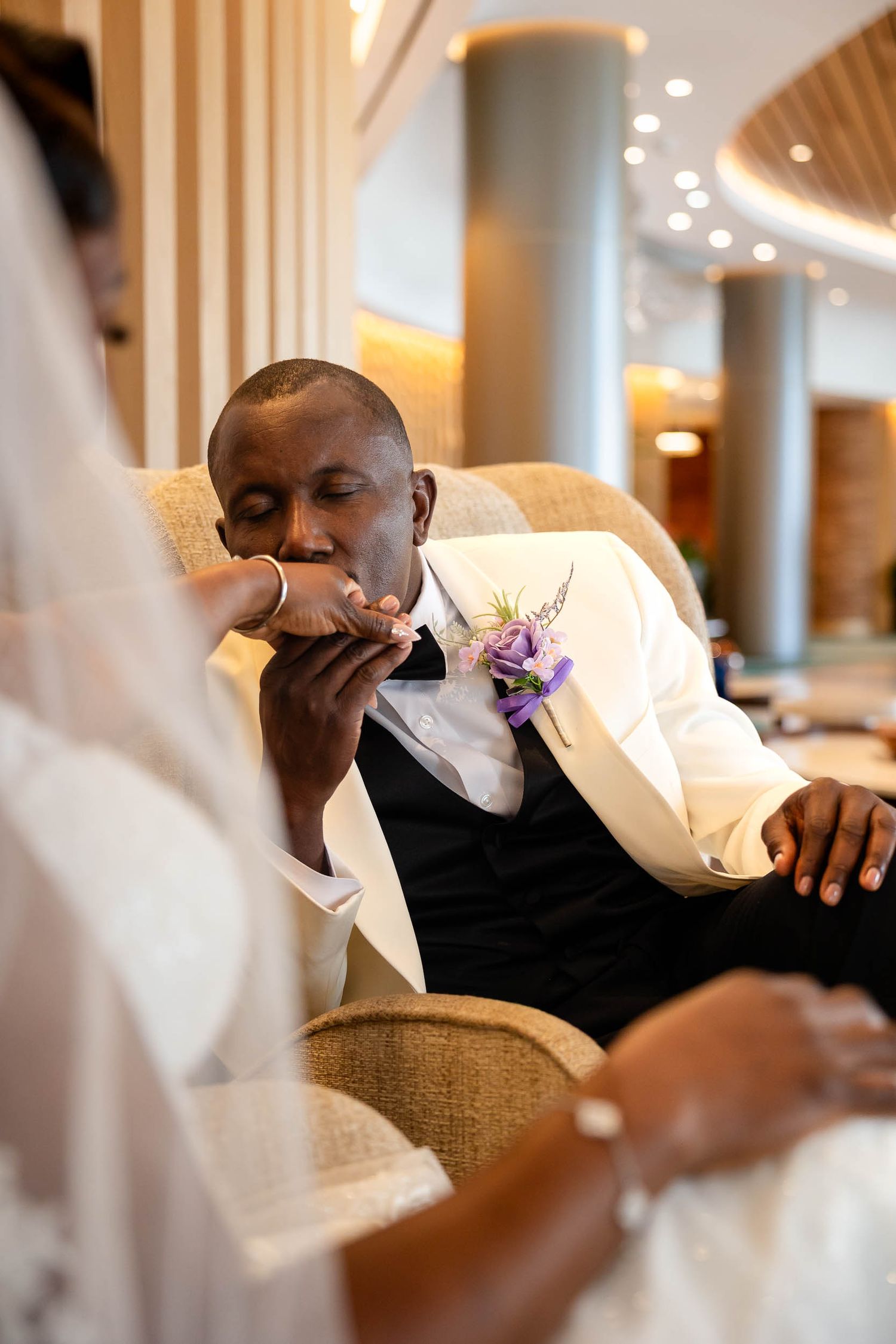 Groom in white tuxedo jacket kisses bride's hand tenderly while seated in elegant chair during wedding.