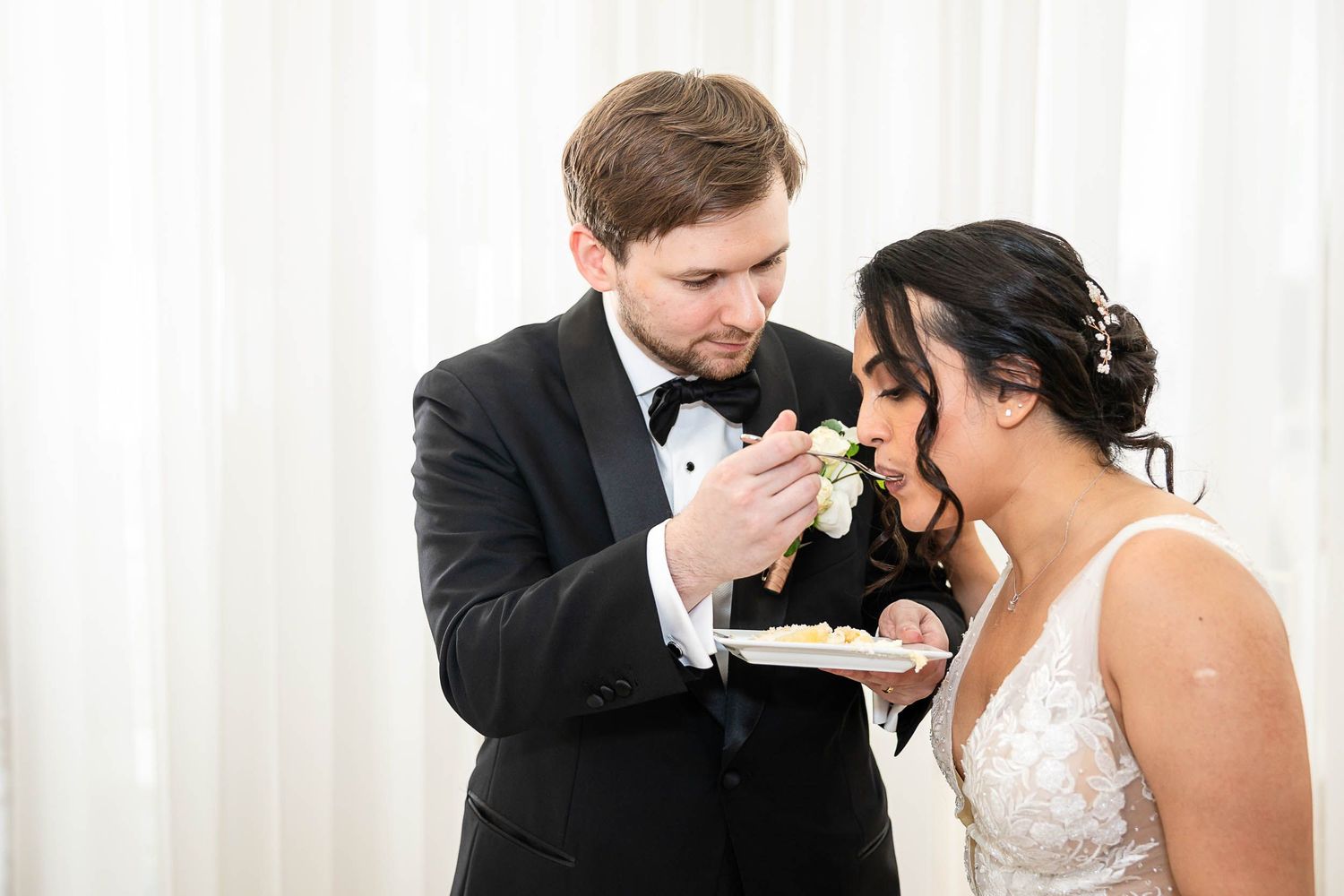 Groom in tuxedo feeding wedding cake to bride in lace dress during elegant wedding reception.