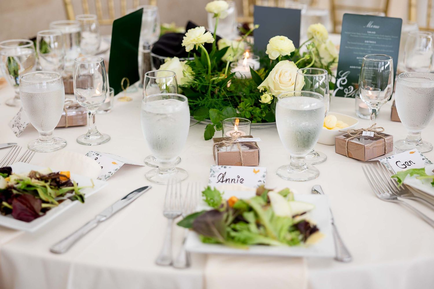 Elegant wedding reception table with white floral centerpiece, place cards, candles, and salad plates on white linen.