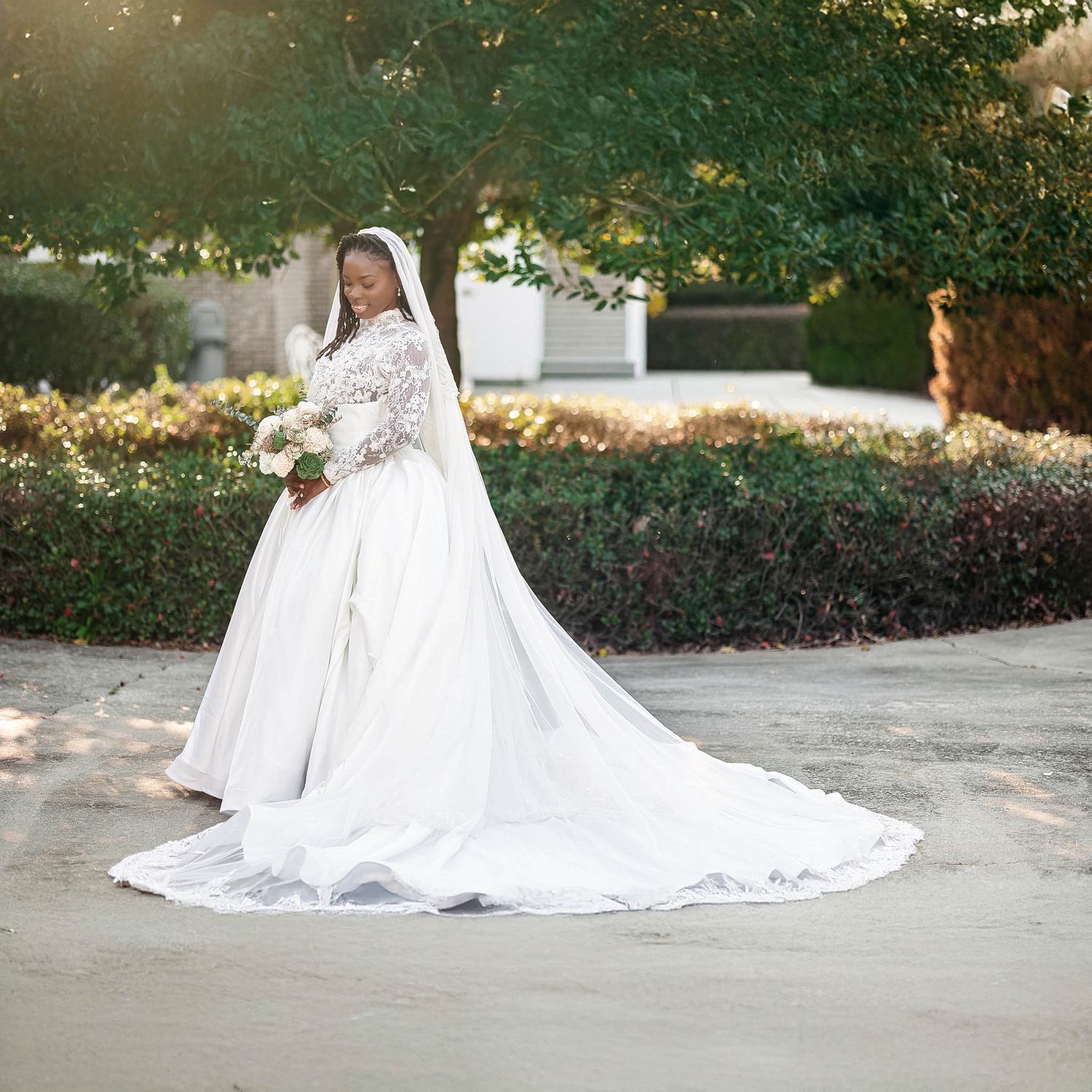 Bride in elegant white lace long-sleeve wedding gown with cathedral train, holding bouquet outdoors.