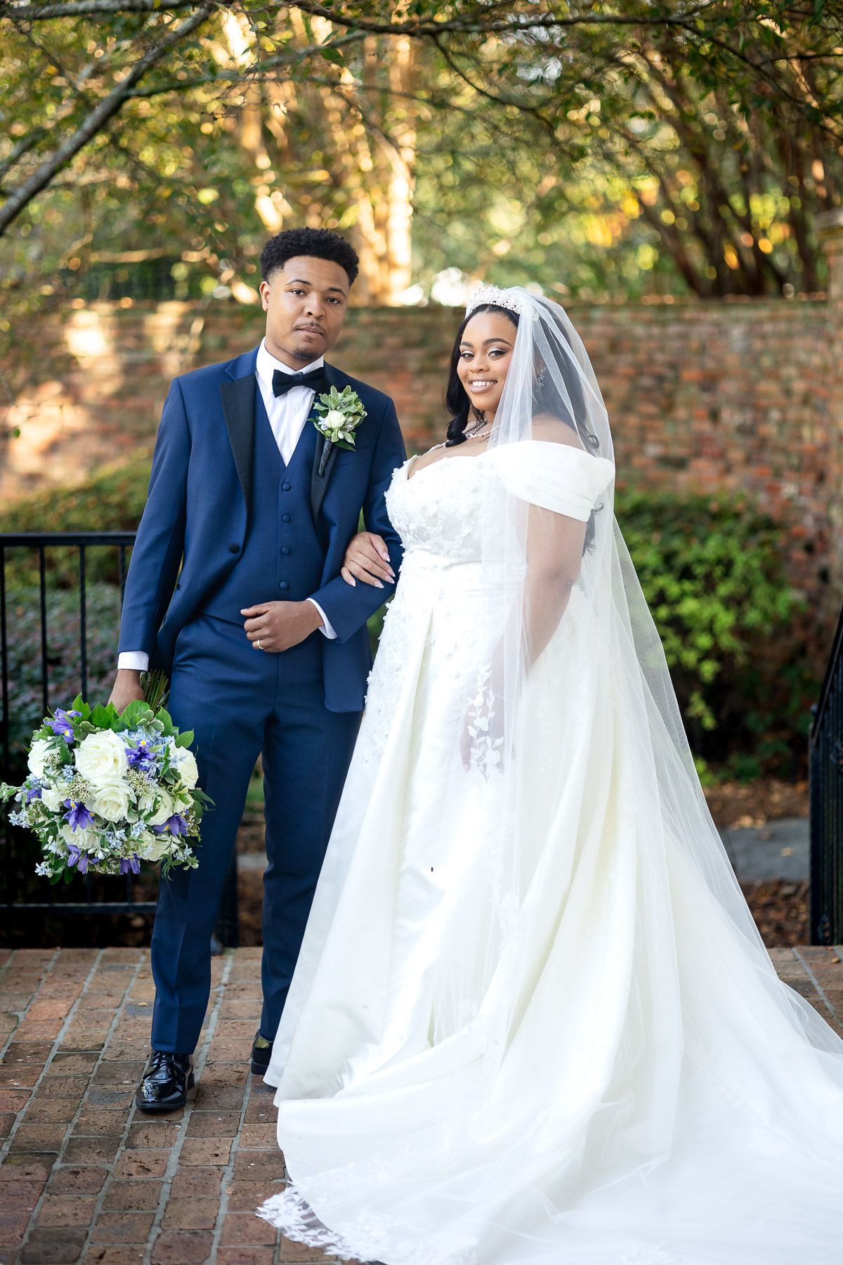 A bride in a white off-shoulder gown and veil poses with a groom in a navy blue suit holding a bouquet outdoors.