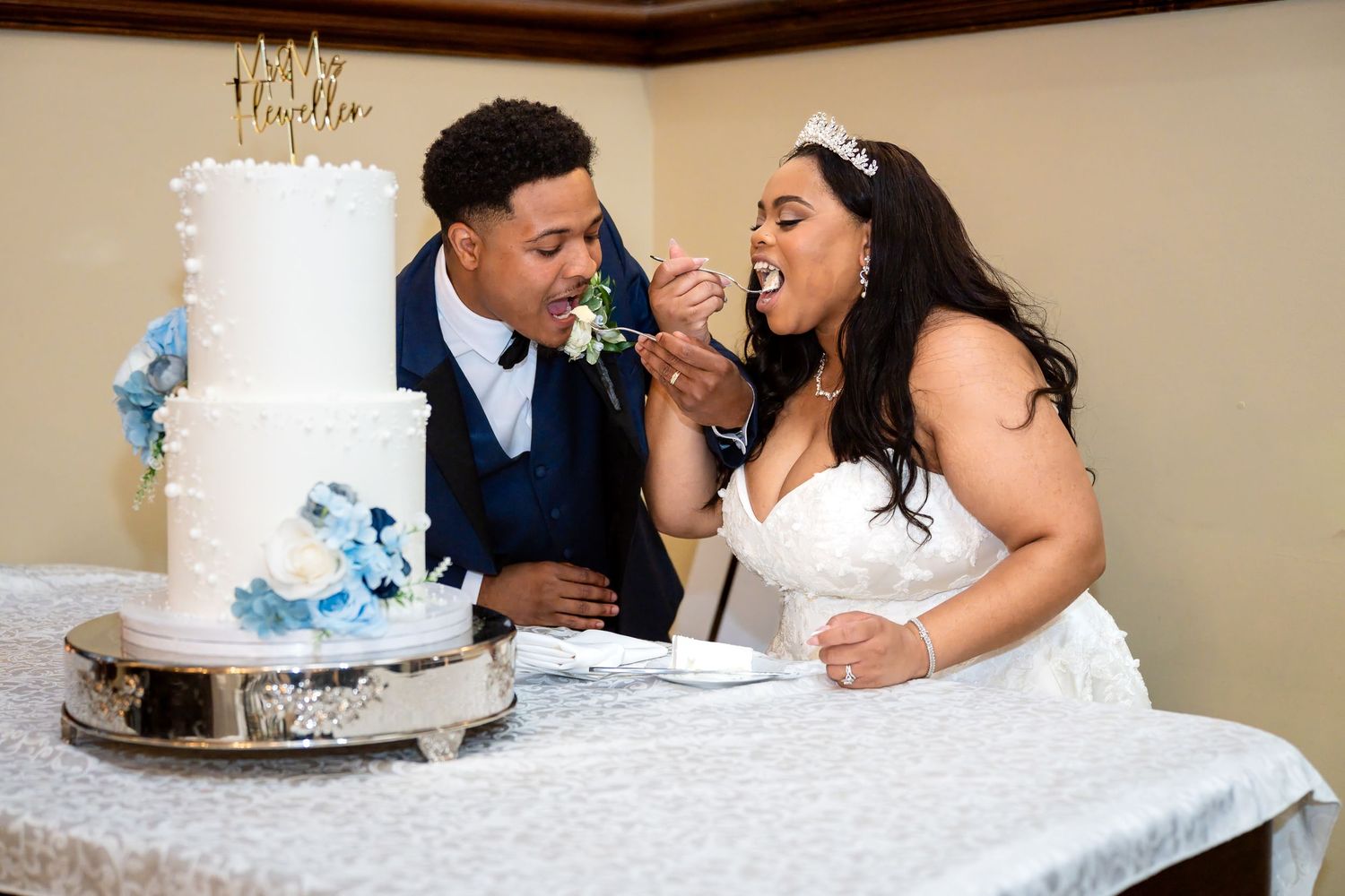 Bride and groom feeding each other wedding cake at reception, white tiered cake with blue floral decorations.