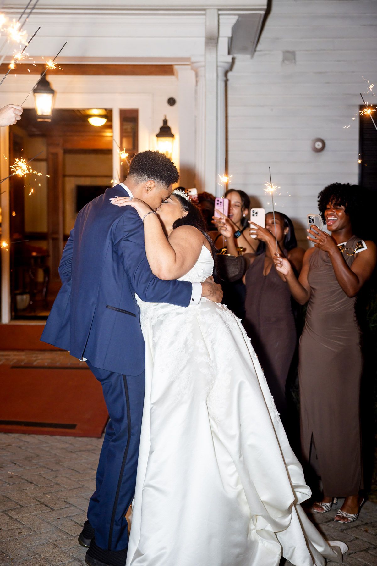 Bride and groom share a romantic kiss during sparkler send-off at outdoor wedding reception with guests cheering.