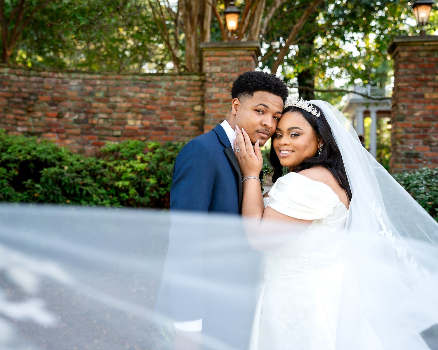 A bride and groom pose romantically outdoors, her white veil flowing, against a brick wall backdrop.