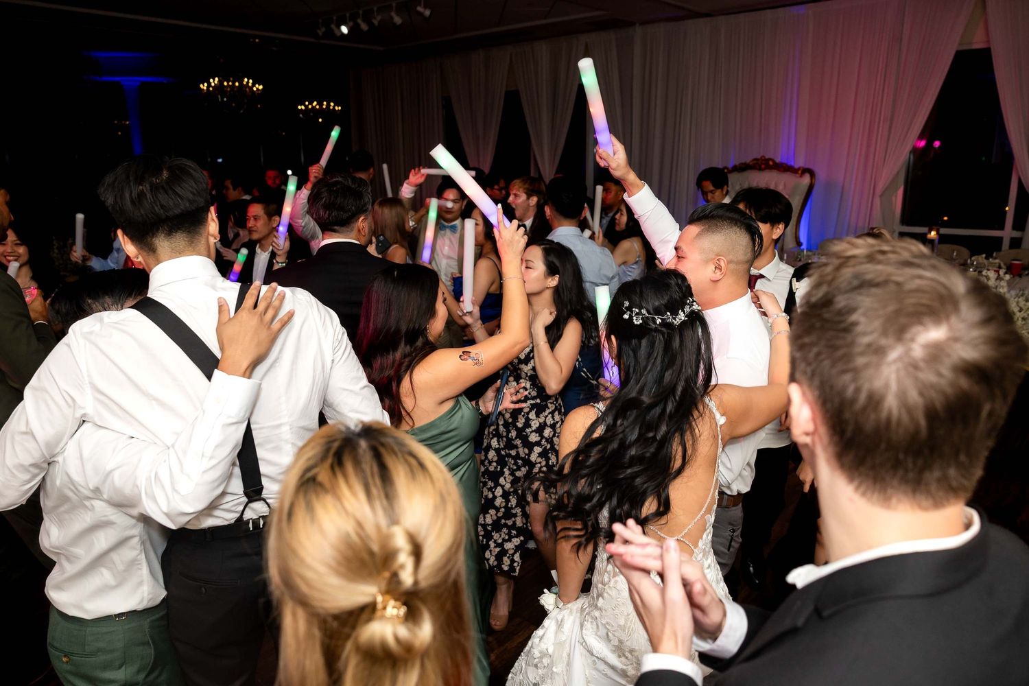 Wedding guests dance and celebrate with glow sticks on packed reception dance floor at night.