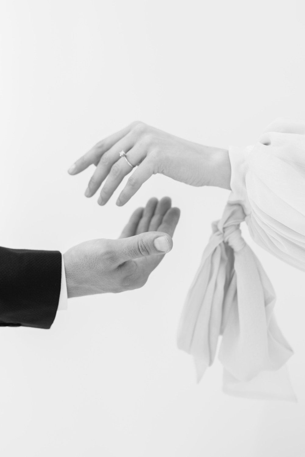 Black and white photo of bride and groom hands, woman wearing wedding ring, man reaching out to hold her hand.