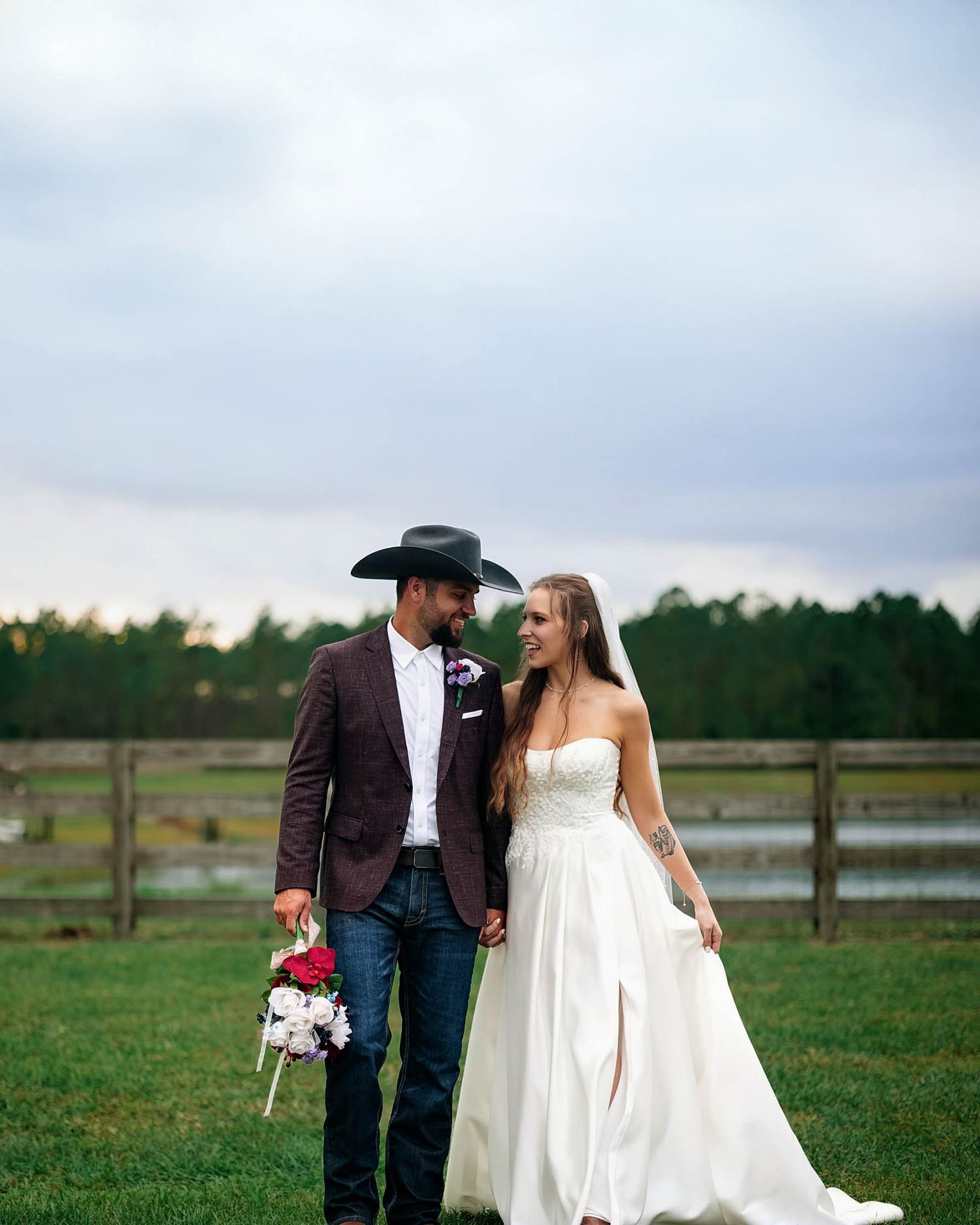Bride in white strapless gown and groom in brown blazer, jeans, and black cowboy hat holding bouquet at outdoor farm wedding.