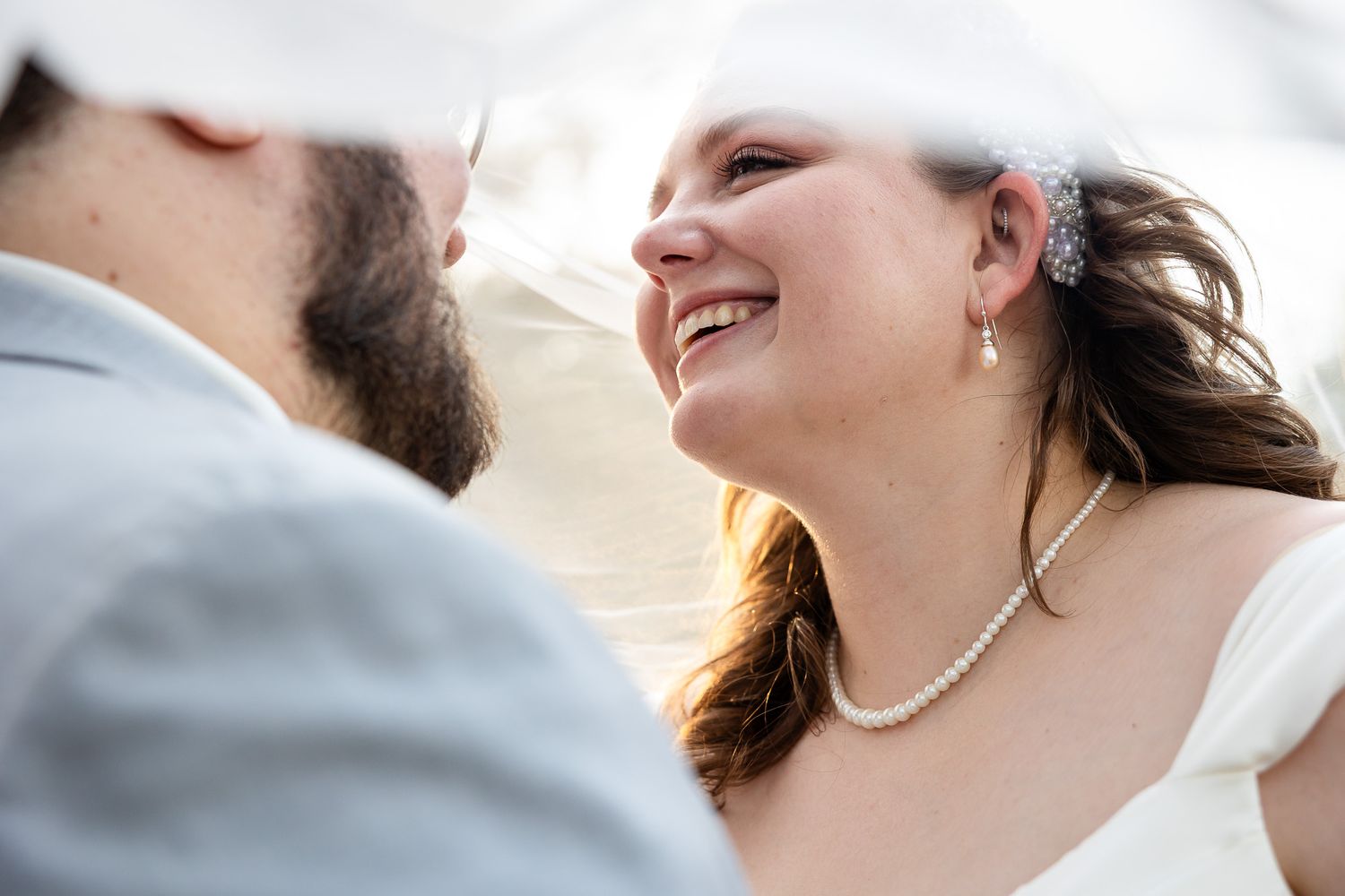 Bride smiling joyfully at groom during outdoor wedding ceremony, wearing pearl necklace and veil with decorative hairpiece.
