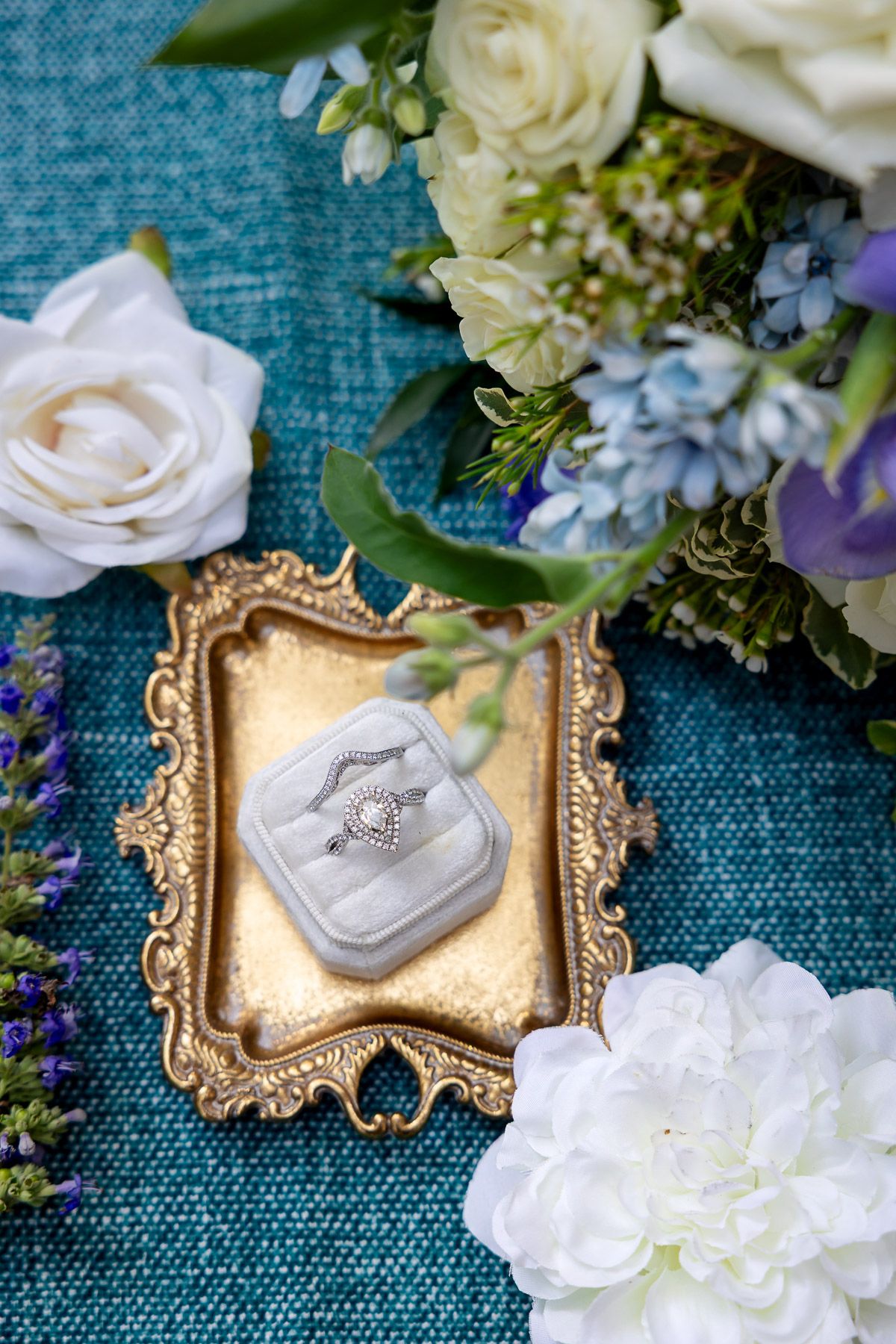 Wedding rings in velvet box on gold tray surrounded by white, blue, and purple flowers on teal background.