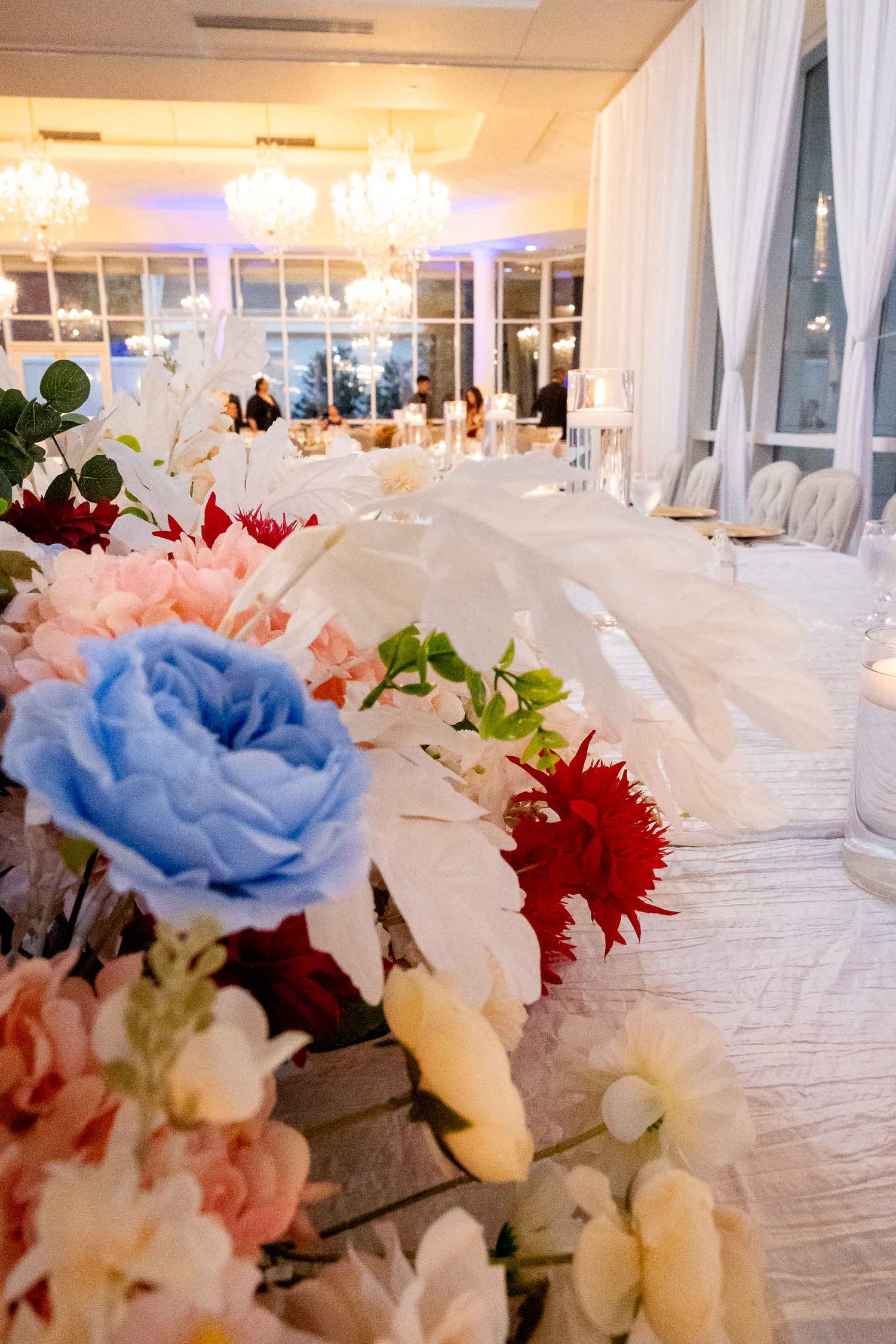 Elegant wedding reception table decorated with colorful flowers and white feathers under chandelier lighting.