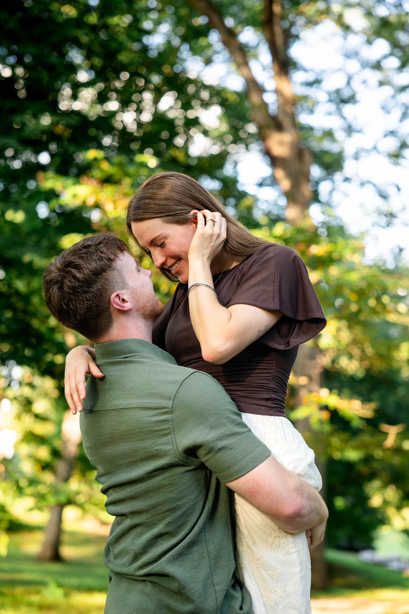 A joyful couple embraces outdoors, the man lifting the laughing woman in a lush green park setting.