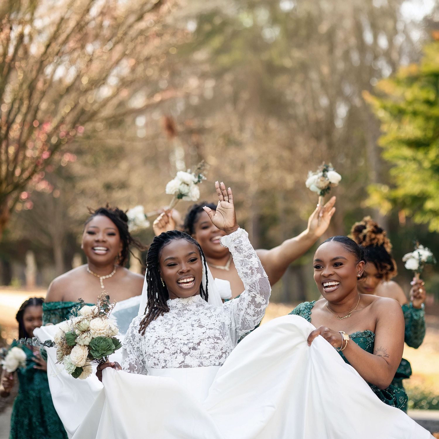 Joyful bride in white lace gown surrounded by bridesmaids in emerald green dresses holding white bouquets outdoors.