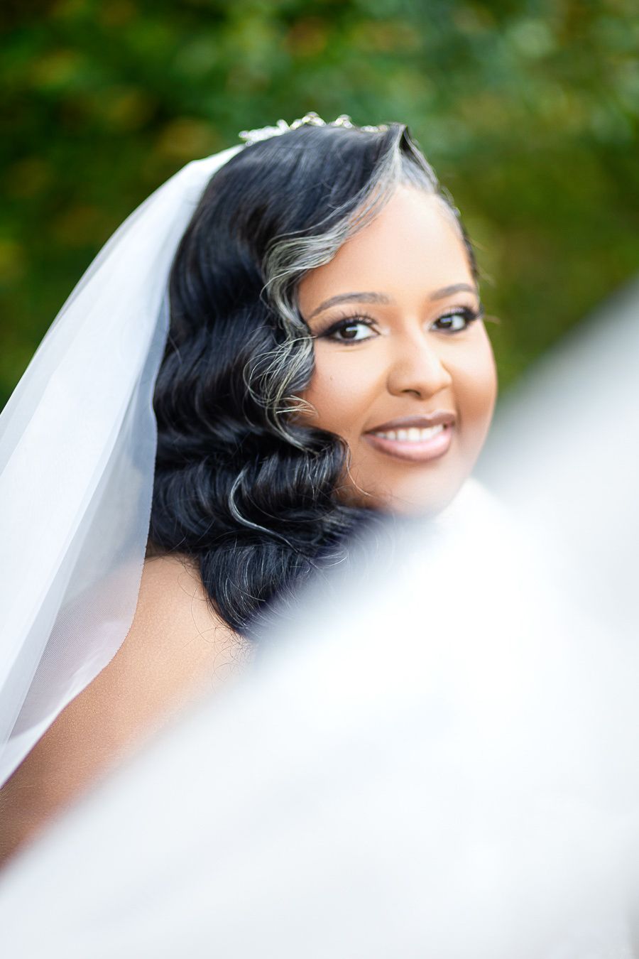 Smiling Black bride wearing a white veil and elegant wavy hairstyle with highlights, posing outdoors on her wedding day.