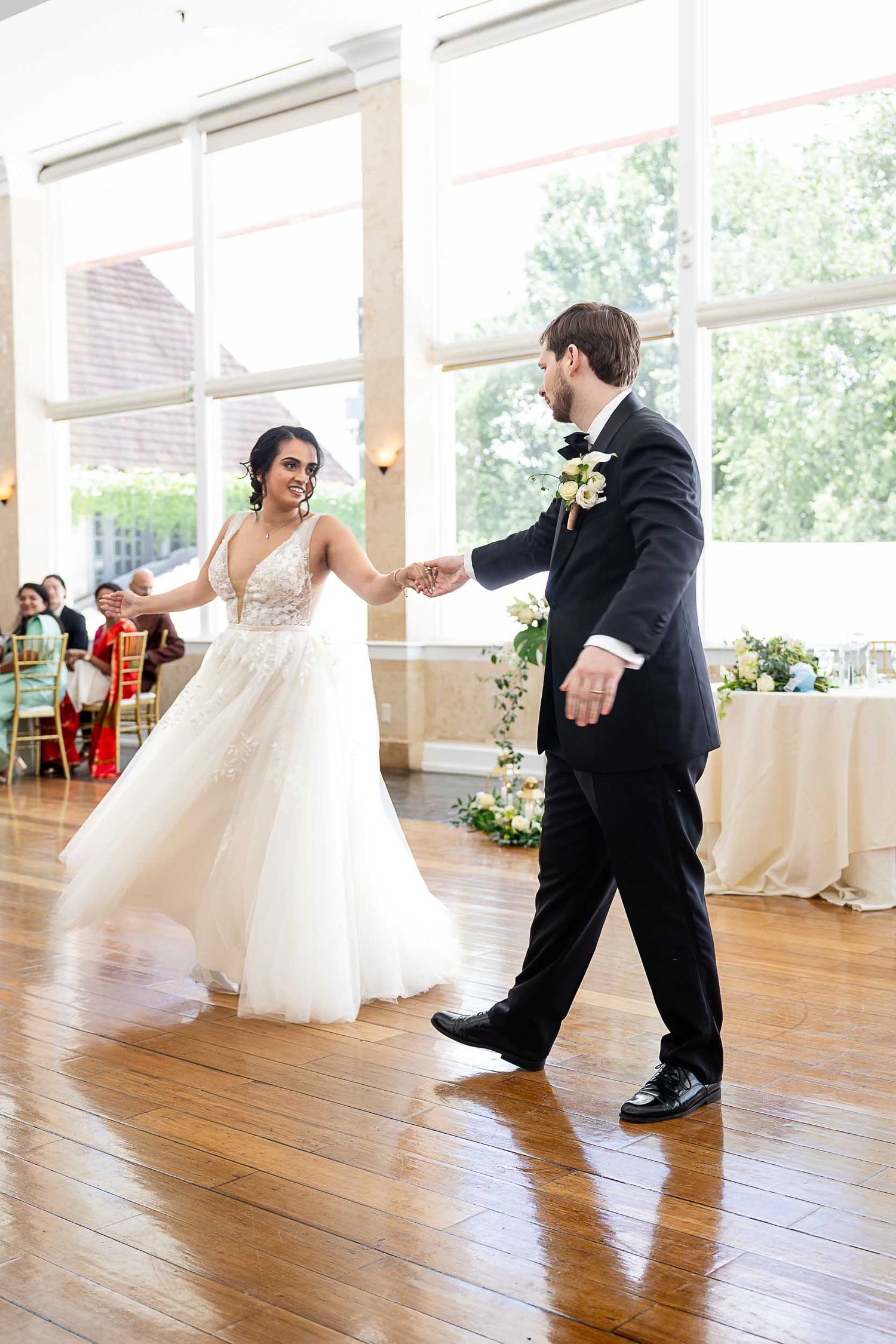 Bride and groom share their first dance, holding hands on a wooden dance floor in a bright, elegant wedding reception venue.