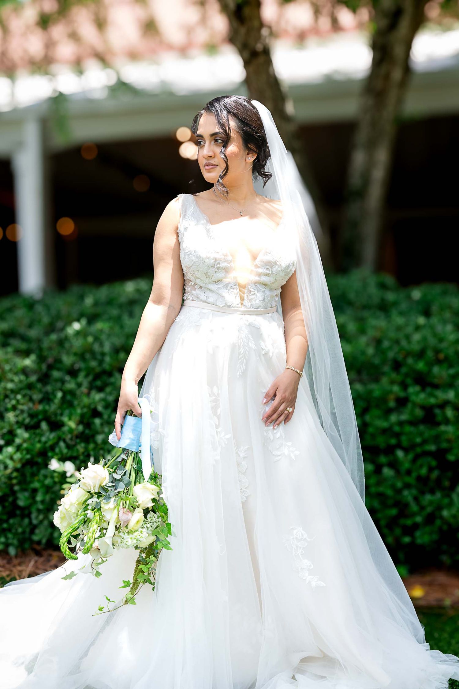 Bride in white lace ballgown with veil holding bouquet of white flowers outdoors.