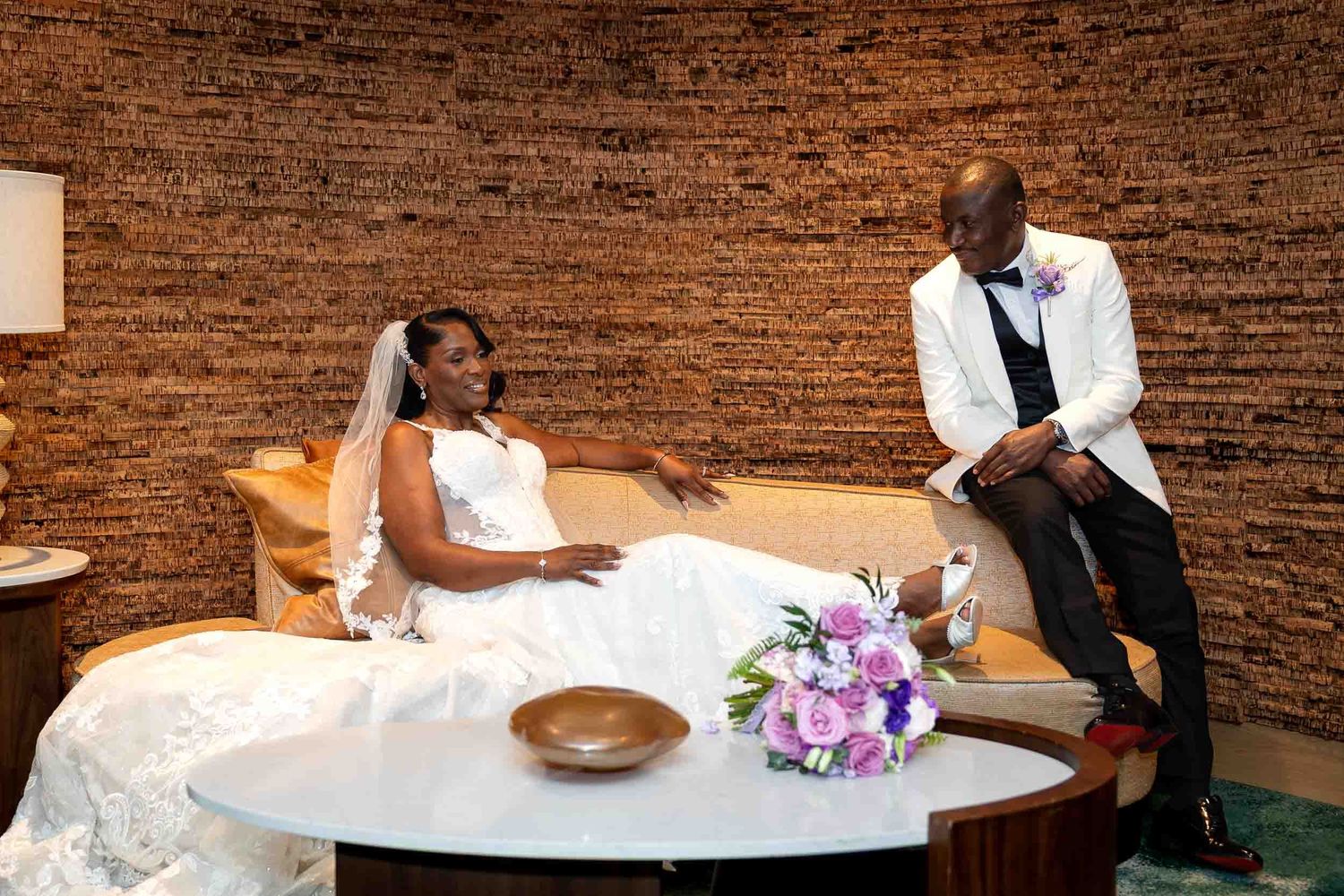 Bride in white gown relaxes on sofa with groom in white blazer at modern wedding venue.