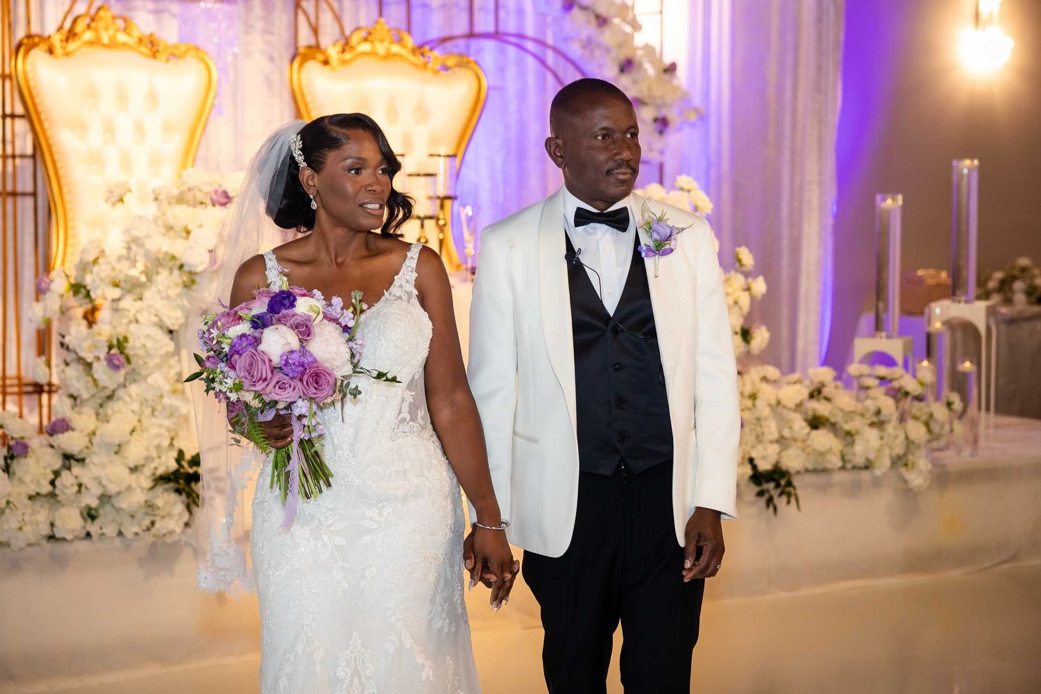 Bride and groom holding hands smiling at elegant wedding reception with purple floral backdrop.