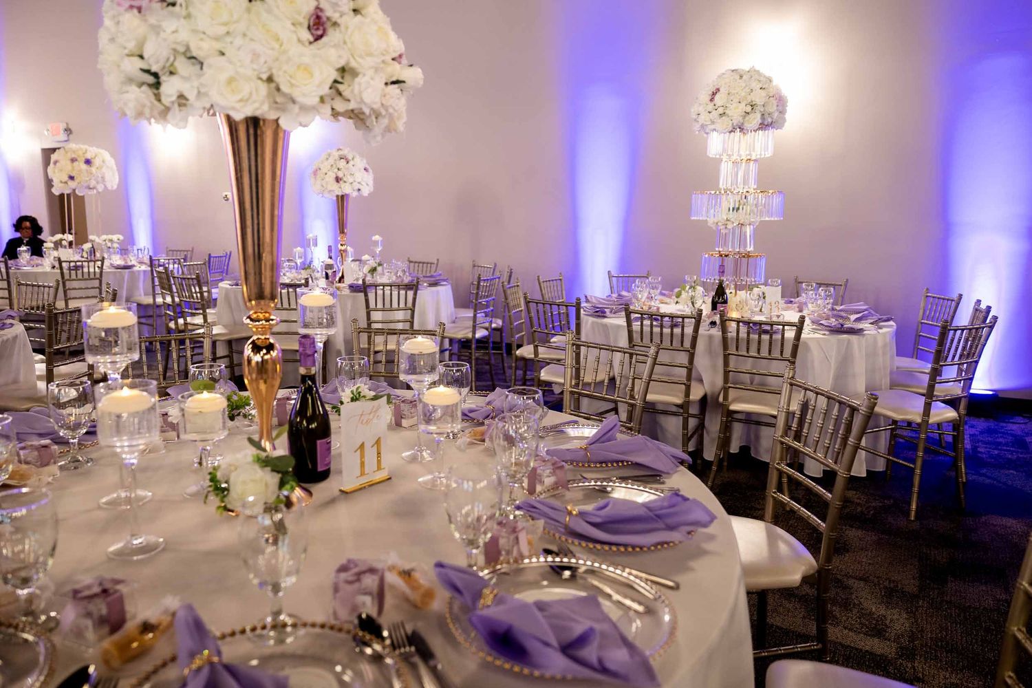 Luxurious wedding reception hall with lavender uplighting, silver chiavari chairs, white floral centerpieces, and purple napkins.