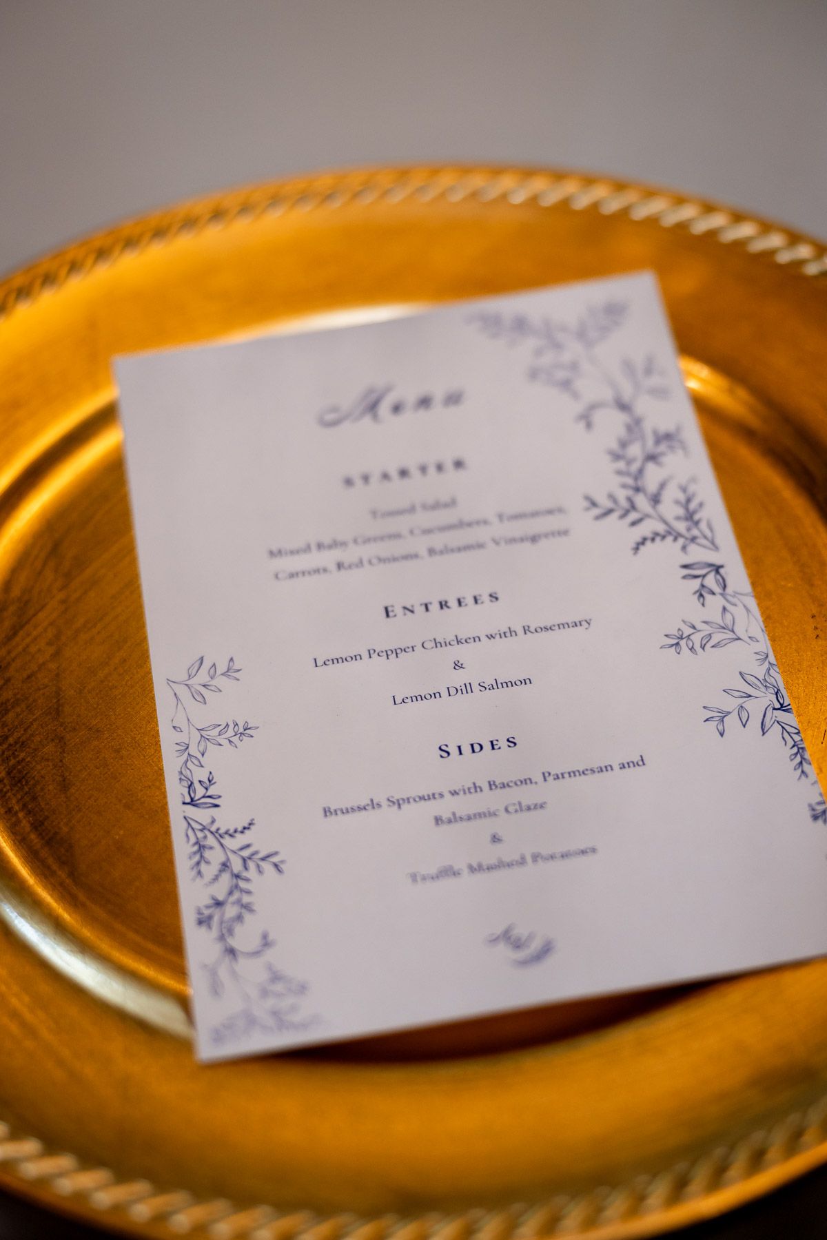 Elegant wedding menu card on gold charger plate listing starter, entrees, and sides with floral border.
