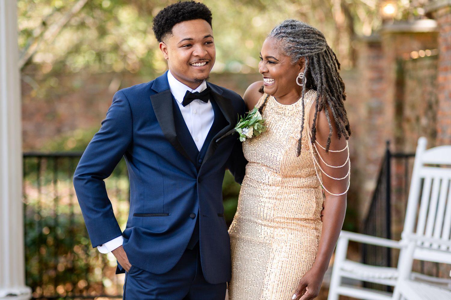 A young man in a navy tuxedo and a woman in a gold gown smile together outdoors at a prom event.