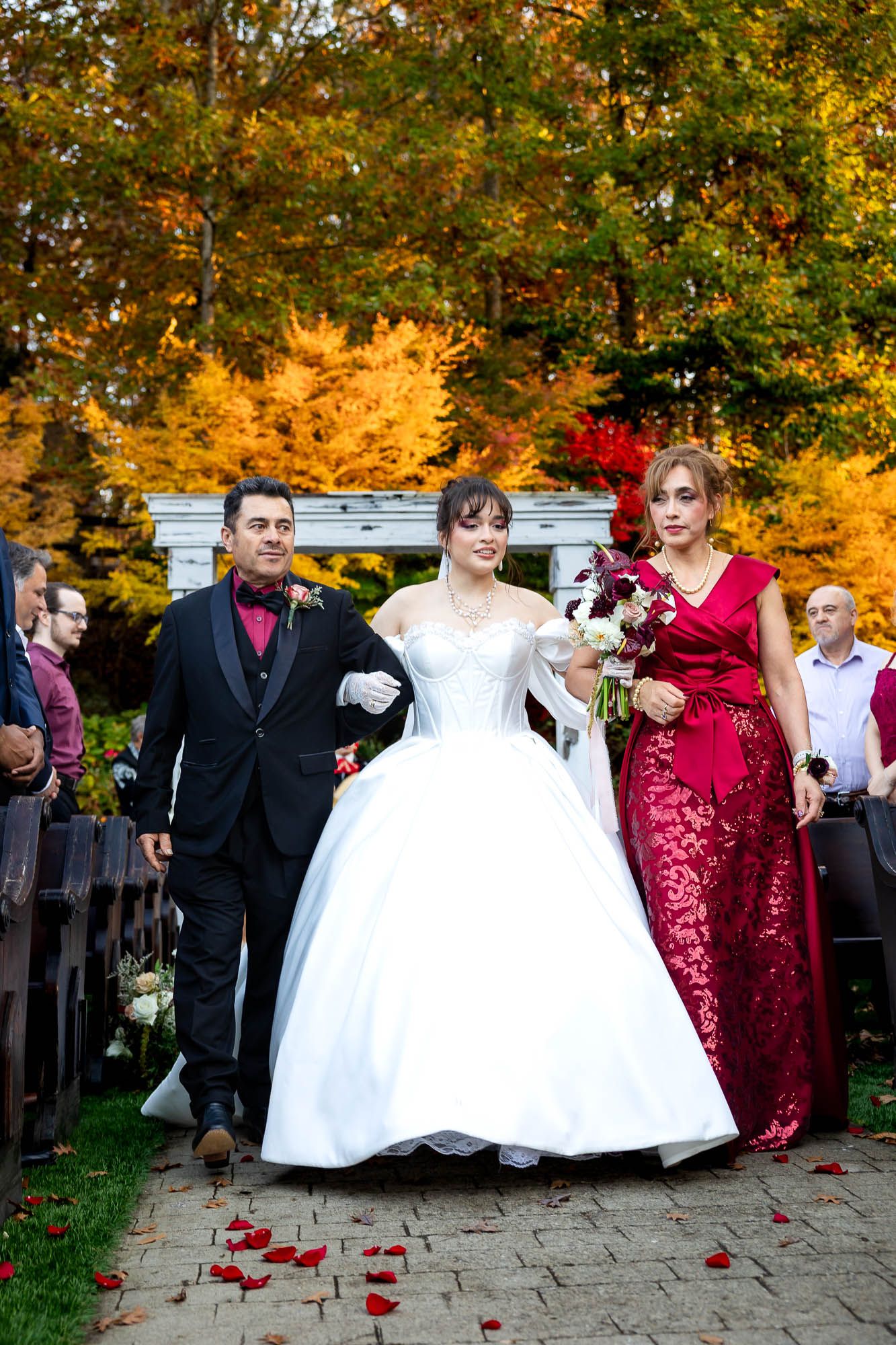 Bride in white gown walks with man in black tuxedo and woman in red dress at outdoor fall wedding ceremony.