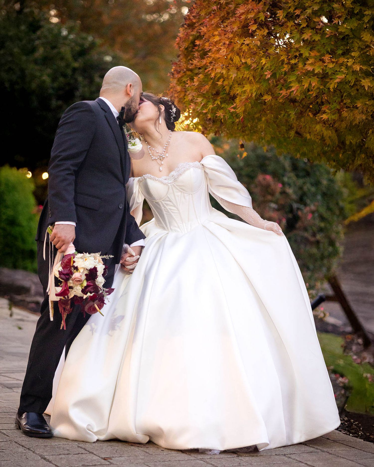 Couple sharing a kiss in autumn garden, bride in white ballgown holding burgundy bouquet.